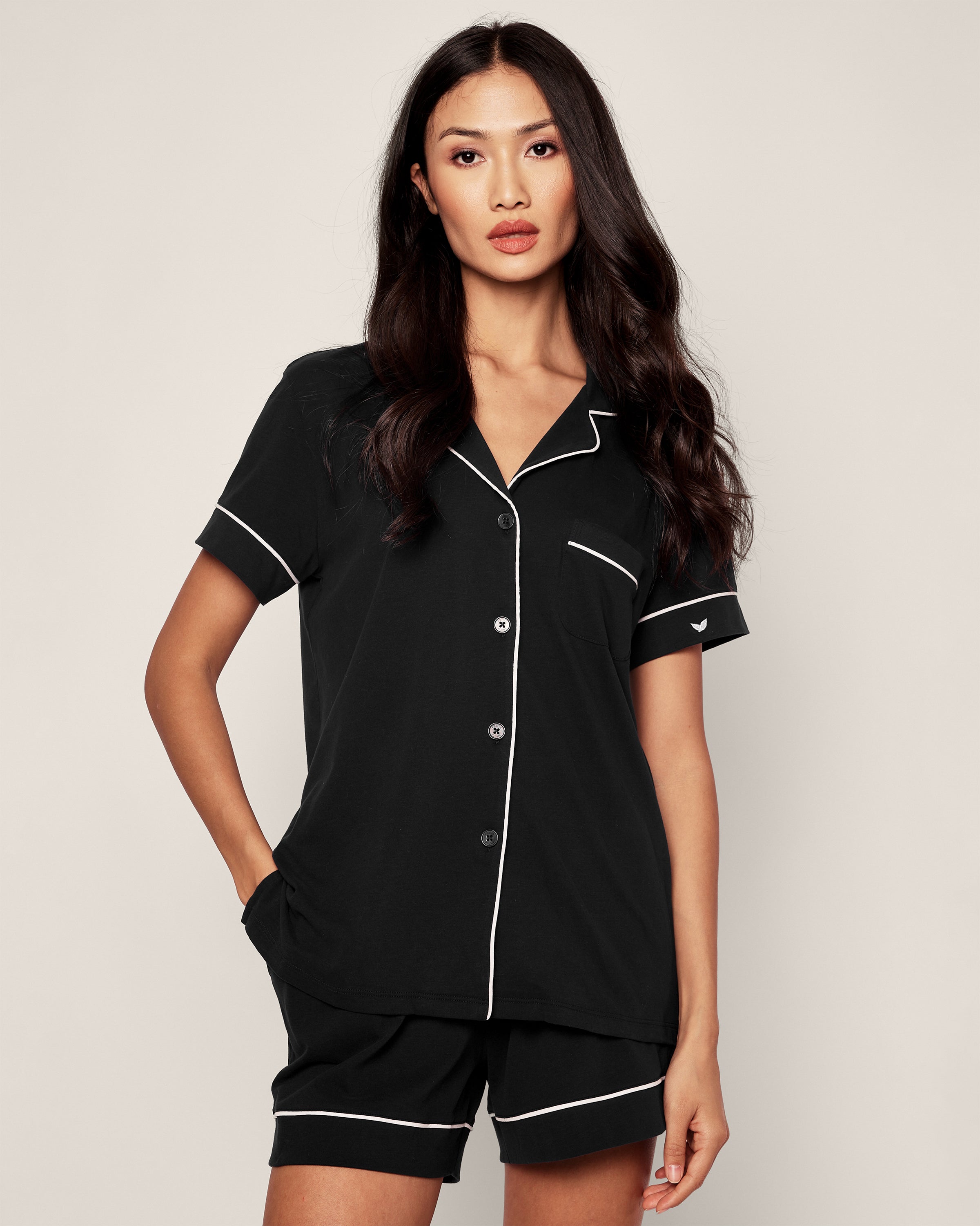 A person models the luxurious Petite Plume Womens Pima Pajama Short Set in Black, featuring white piping. The ensemble includes a short-sleeve buttoned shirt and shorts. They stand against a plain background, hand in pocket, looking at the camera.