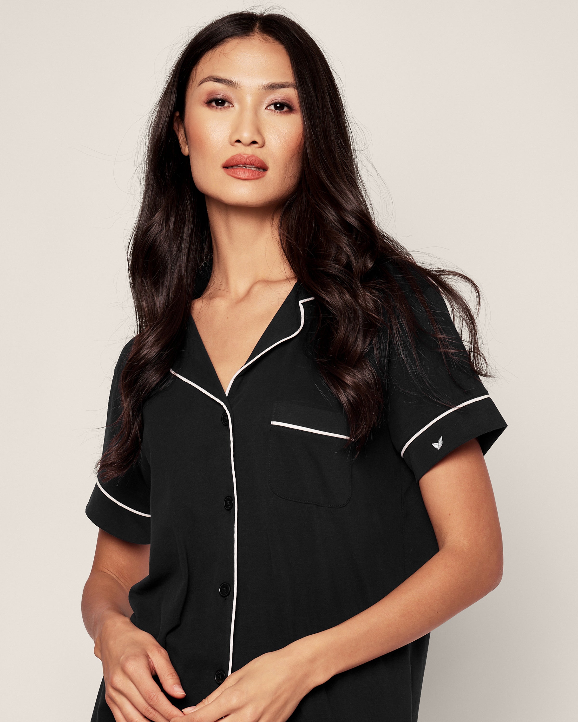 A person with long, wavy hair is wearing the Womens Pima Pajama Short Set in Black by Petite Plume, featuring a black short-sleeved shirt with white piping and buttons. The shirt has a chest pocket with white trim and an embroidered sleeve detail. The background is plain.