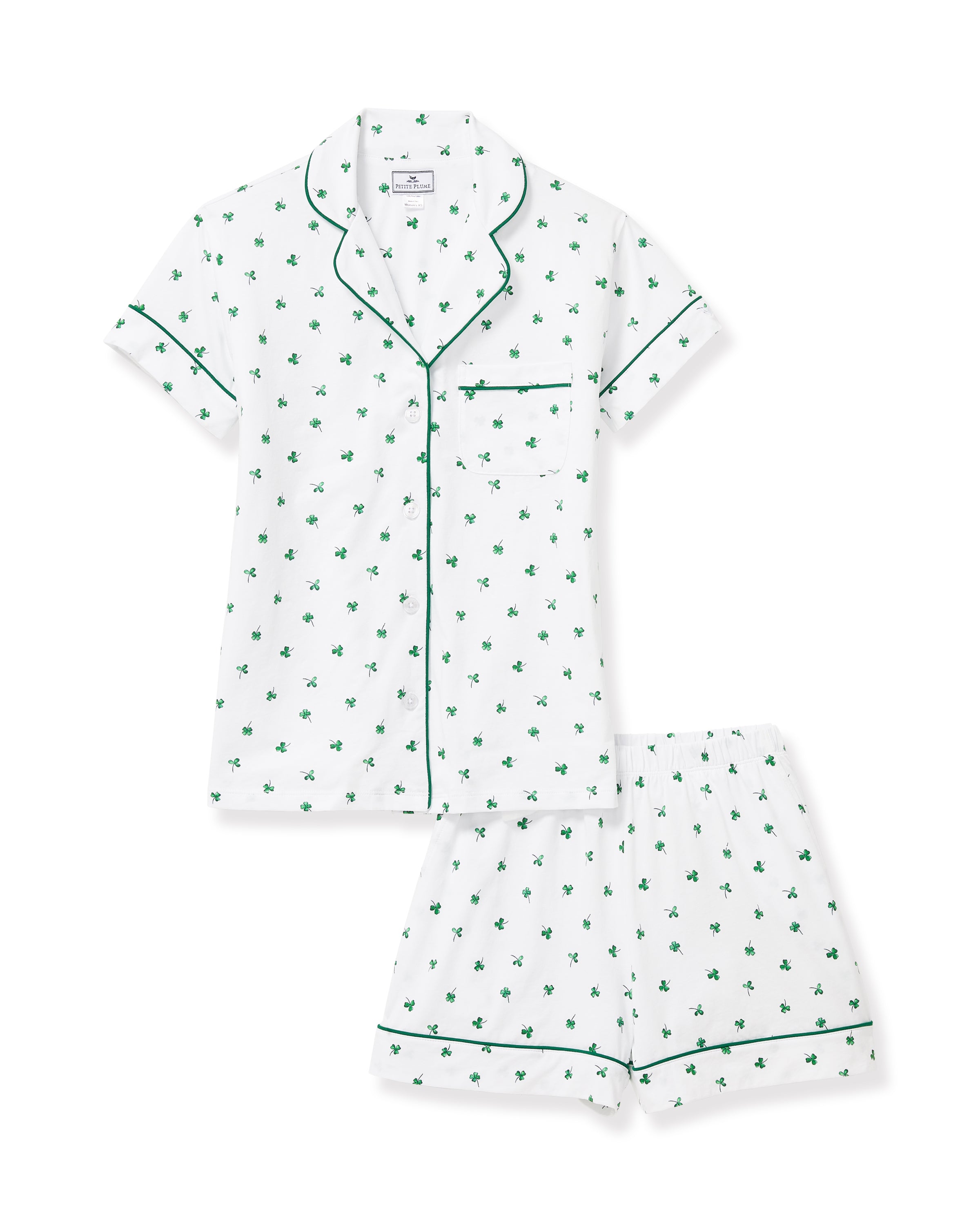 A woman with wavy blonde hair wears the Women's Pima Short Sleeve Short Set in Shamrocks—white pajamas with green clover prints, green trim, short-sleeve top and pocket, plus ruffled-hem shorts—standing against a plain background.