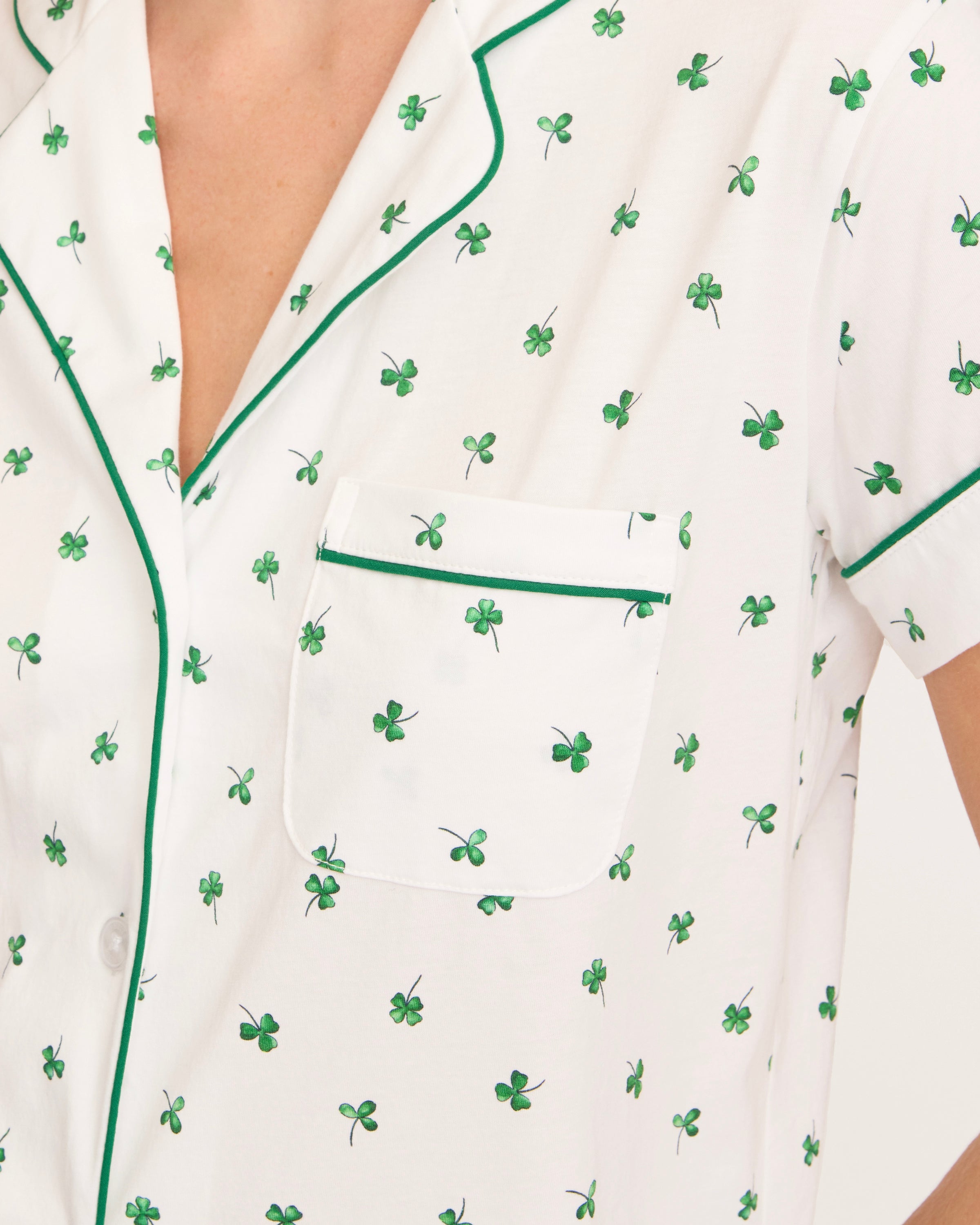 A woman with wavy blonde hair wears the Women's Pima Short Sleeve Short Set in Shamrocks—white pajamas with green clover prints, green trim, short-sleeve top and pocket, plus ruffled-hem shorts—standing against a plain background.
