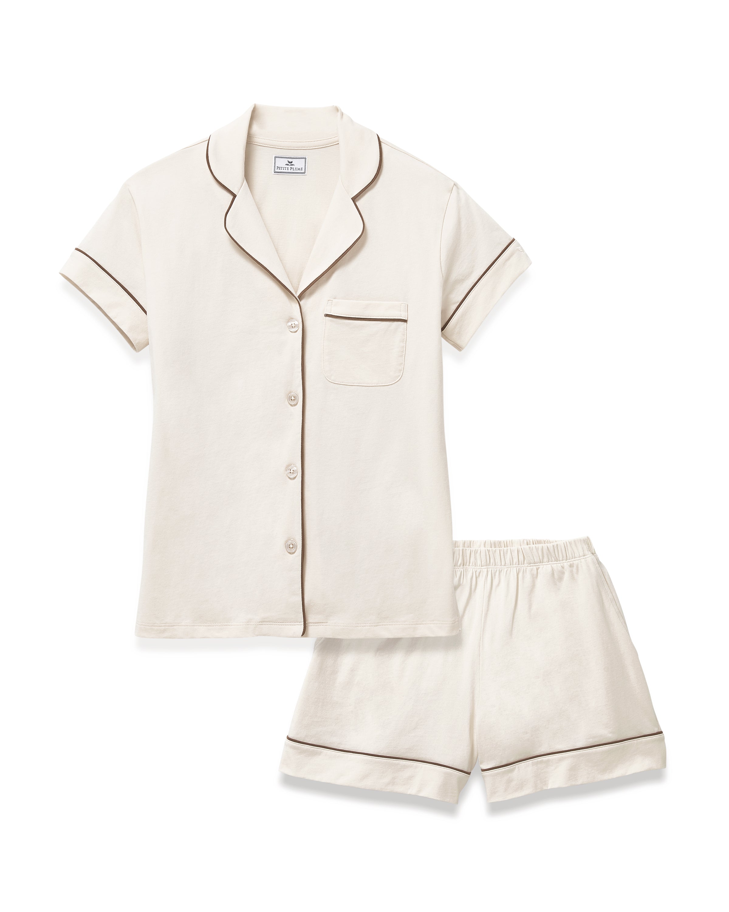 A woman with long curly hair models the Women's Lightweight Pima Short Sleeve Short Set in Ivory, featuring a button-up top with piping and matching shorts, standing barefoot against a plain light background.
