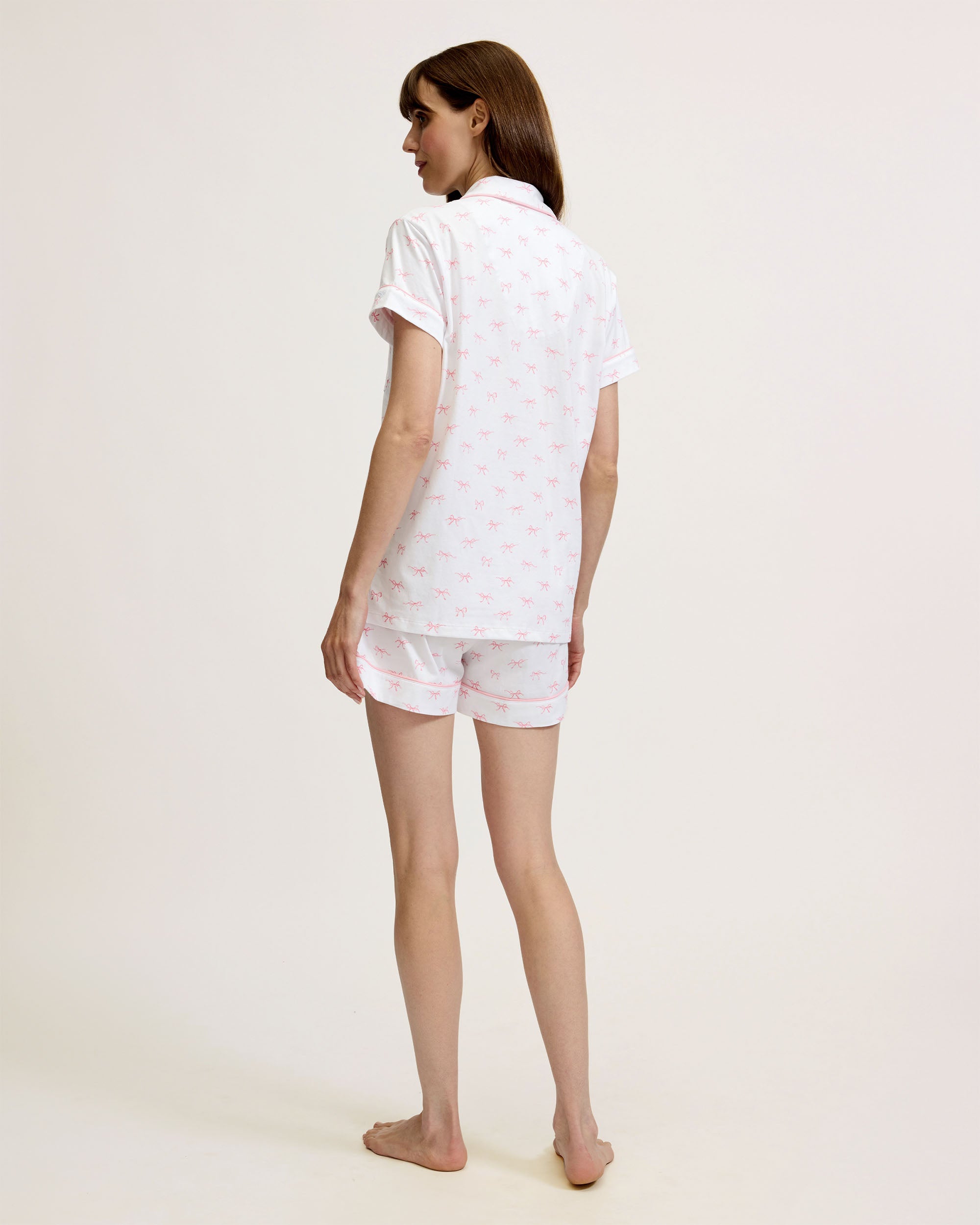 The Women's Pima Short Sleeve Short Set in Blushing Bows features a white button-up top with pink piping, a front pocket, and matching shorts, all adorned with an all-over pink bow pattern.