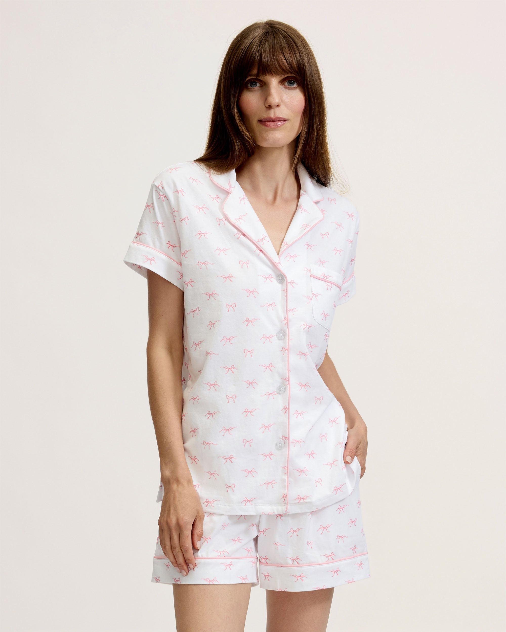 The Women's Pima Short Sleeve Short Set in Blushing Bows features a white button-up top with pink piping, a front pocket, and matching shorts, all adorned with an all-over pink bow pattern.