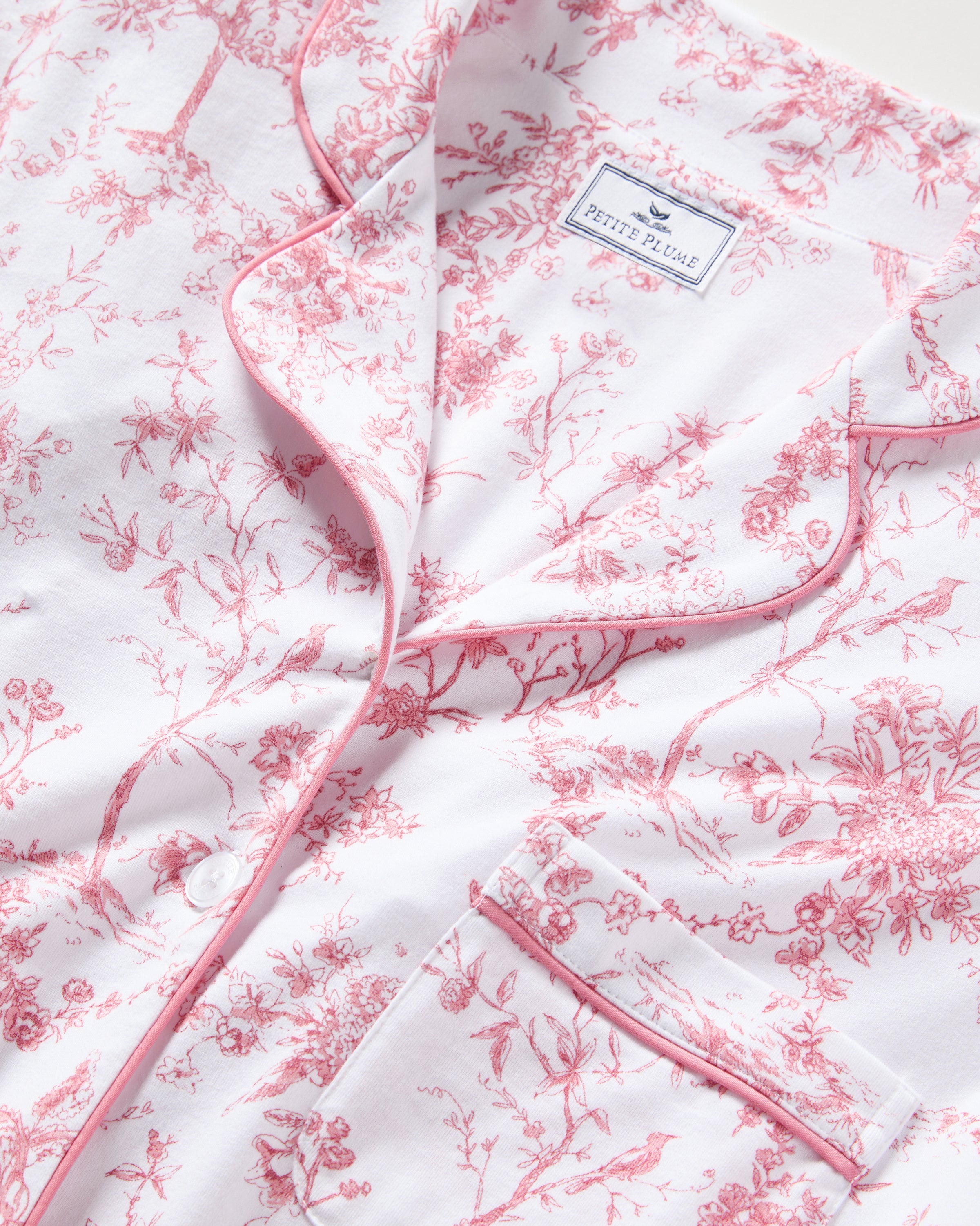 A close-up of the Womens Pima Short Sleeve Short Set in Pink Timeless Toile by Petite Plume showcases botanical elegance with a pink floral pattern and pink piping on the collar and pocket edges.