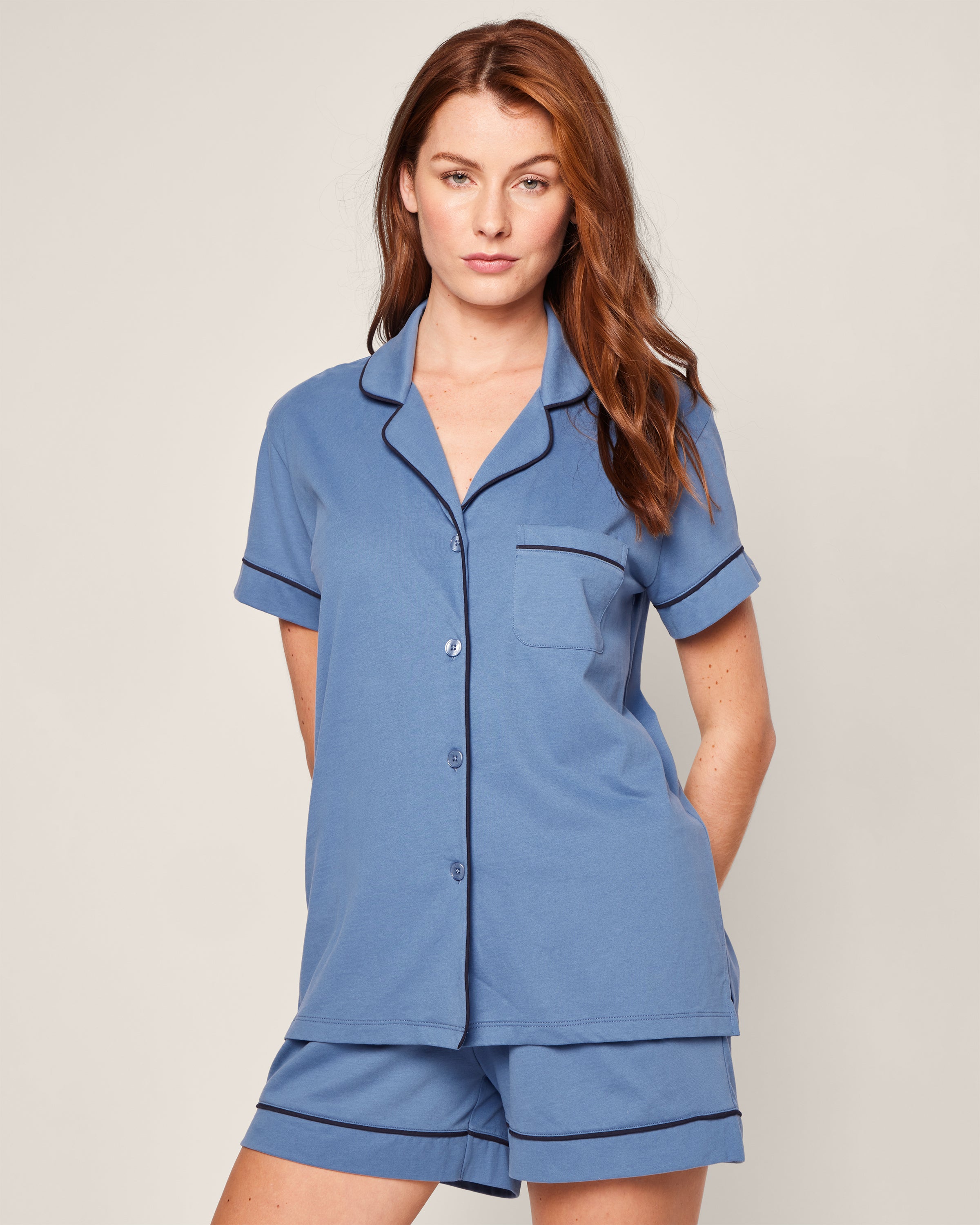 Against a plain backdrop, a person models the Womens Pima Pajama Short Set in Mystique Blue by Petite Plume, with hands discreetly behind their back. The pajamas are made from luxurious Pima cotton featuring elegant black piping on the short-sleeved top and a chest pocket.