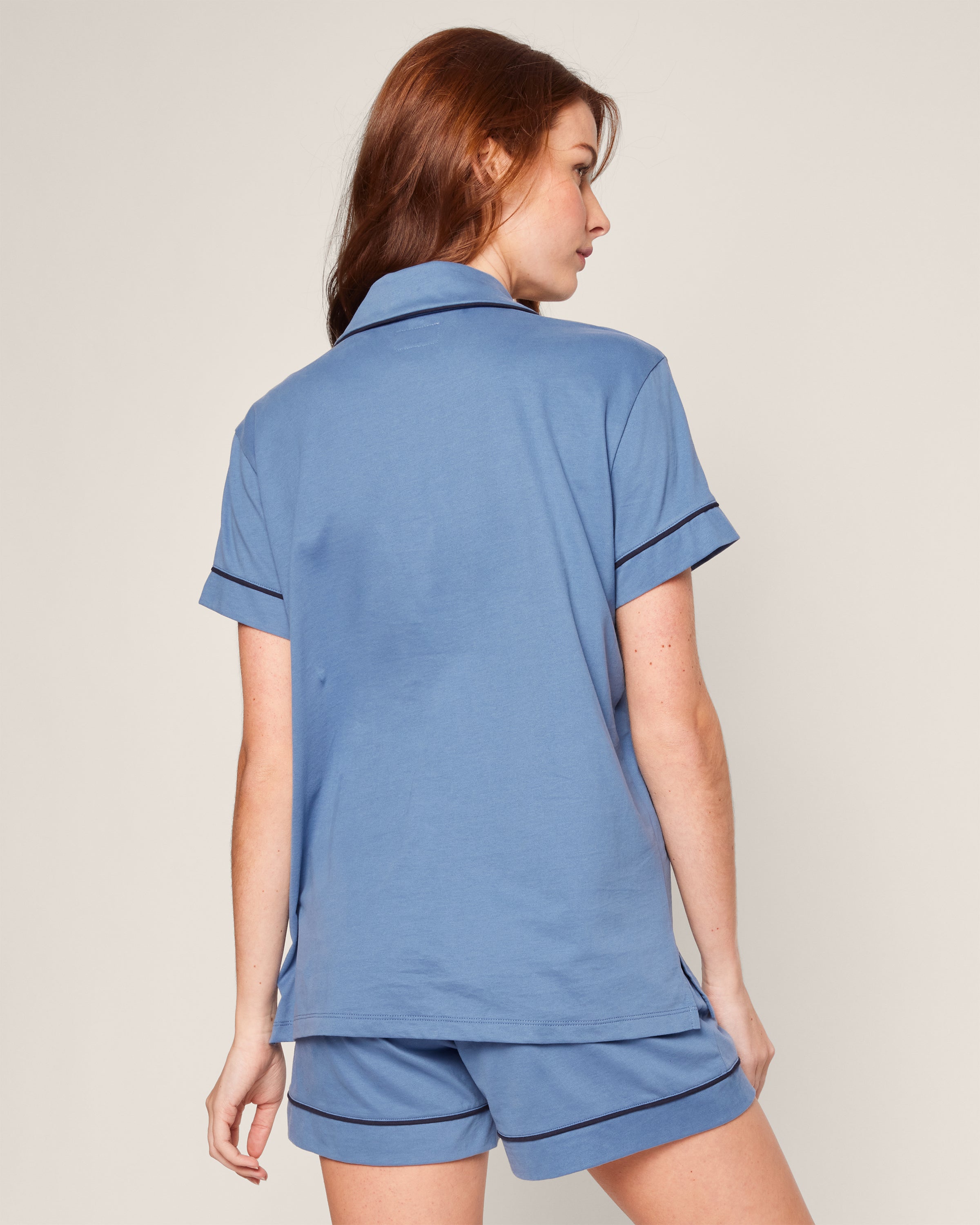 A woman with long red hair, seen from the back, wears Petite Plumes Womens Pima Pajama Short Set in Mystique Blue. The short-sleeved top and matching shorts have dark trim. She is set against a plain, light background.