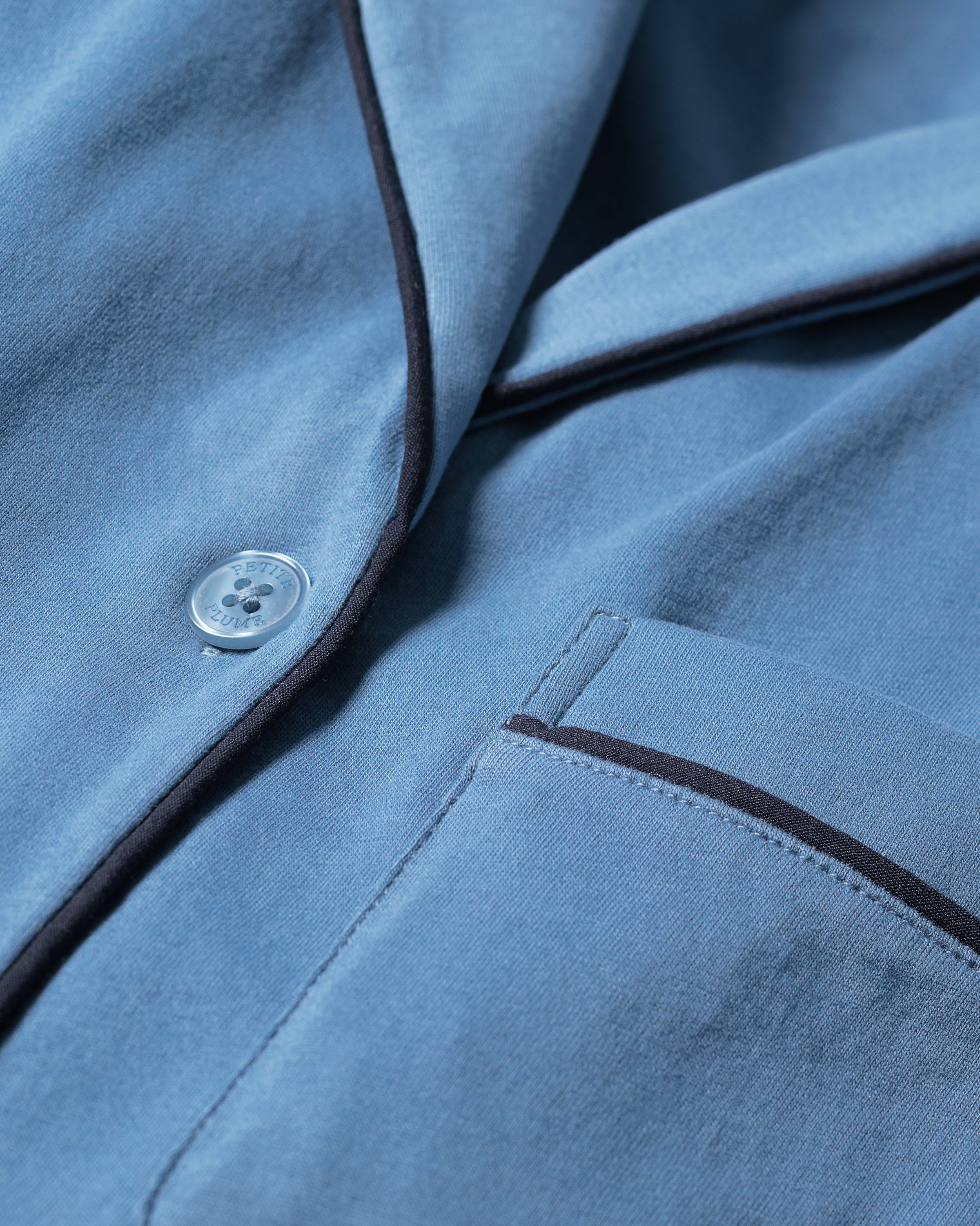A close-up of the Womens Pima Pajama Short Set in Mystique Blue by Petite Plume highlights its serene blue hue, crafted from soft Pima cotton with a button, dark piping along the edges, and a pocket adorned with matching trim. It epitomizes luxury sleepwear.