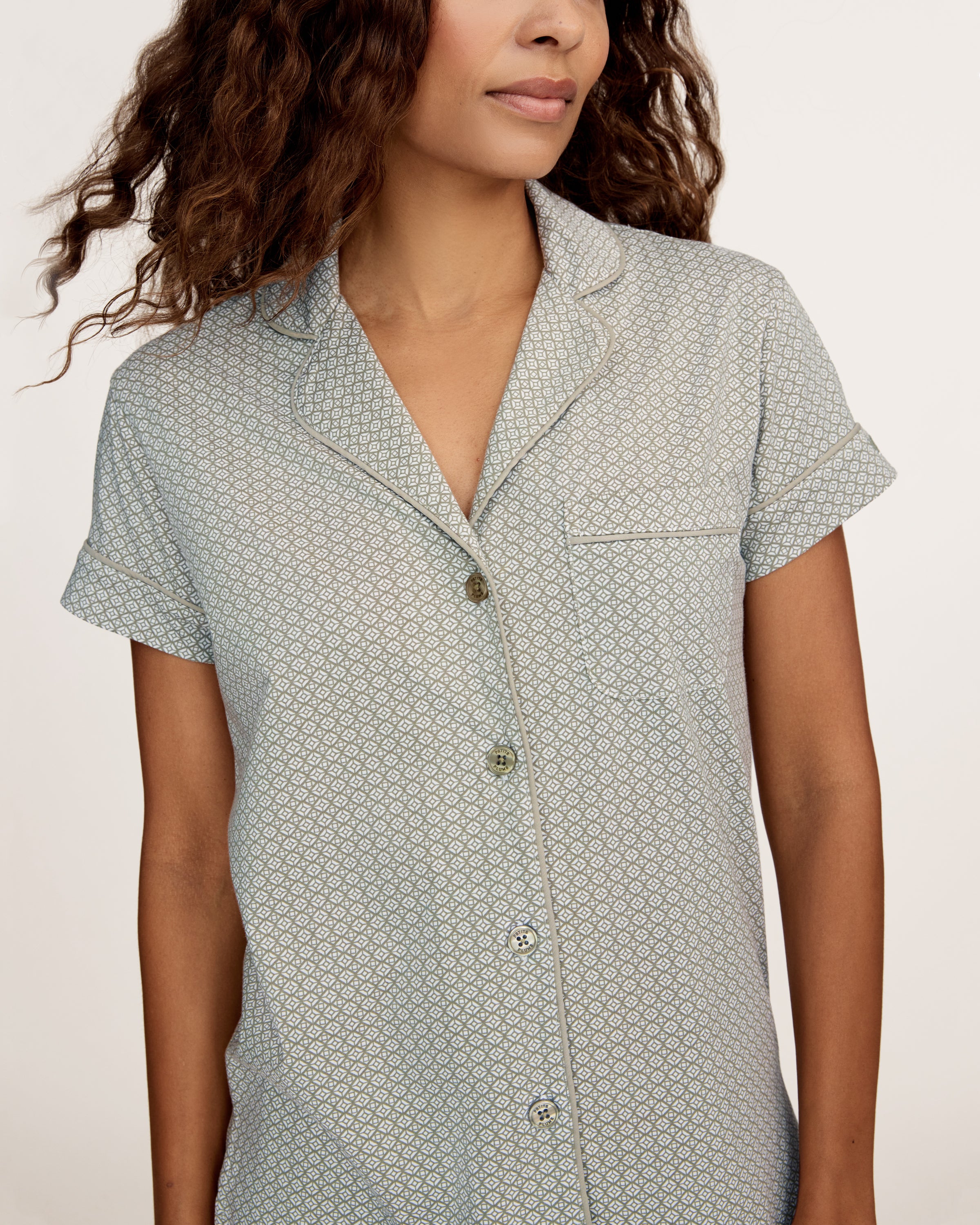 A woman stands barefoot in the Women's Lightweight Pima Short Sleeve Short Set in Sage Geo—featuring a button-up shirt and matching shorts with a small geometric pattern—against a plain light background.