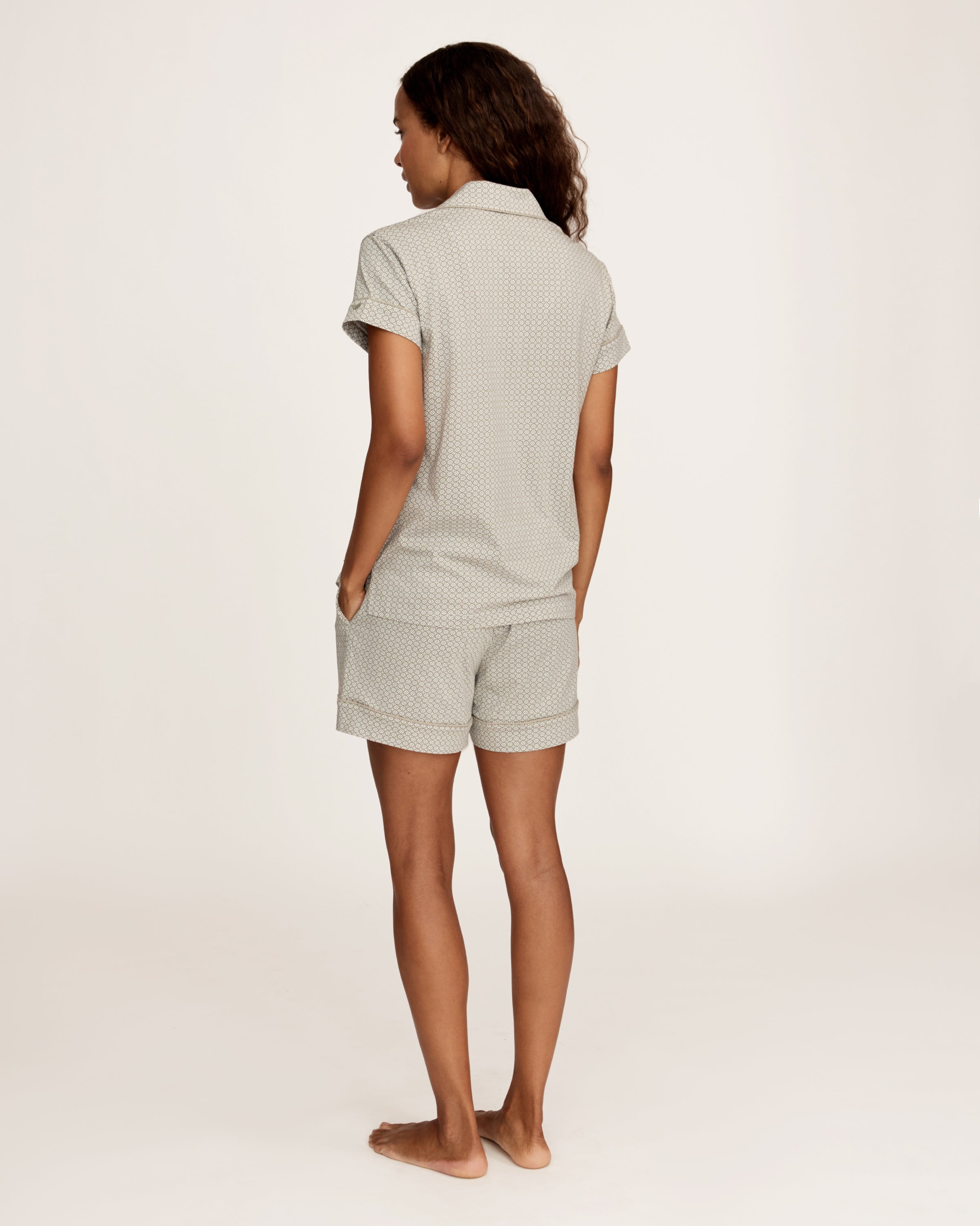 A woman stands barefoot in the Women's Lightweight Pima Short Sleeve Short Set in Sage Geo—featuring a button-up shirt and matching shorts with a small geometric pattern—against a plain light background.