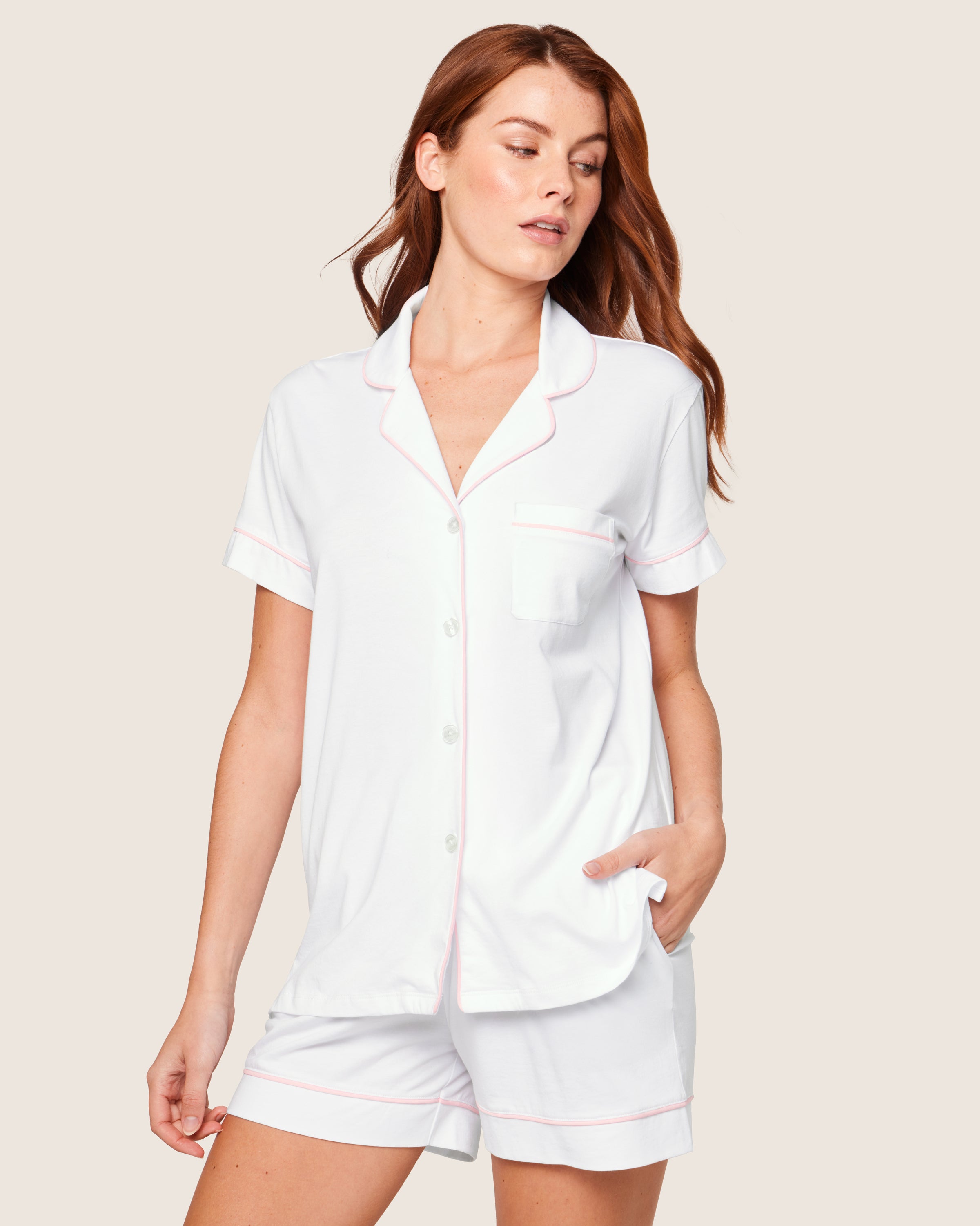 A person with long hair is wearing the Womens Pima Pajama Short Set by Petite Plume in white with pink piping. The top features short sleeves, a button-down front, and a pocket, while the shorts offer a relaxed fit. The neutral background emphasizes the sets luxurious comfort and elegance.
