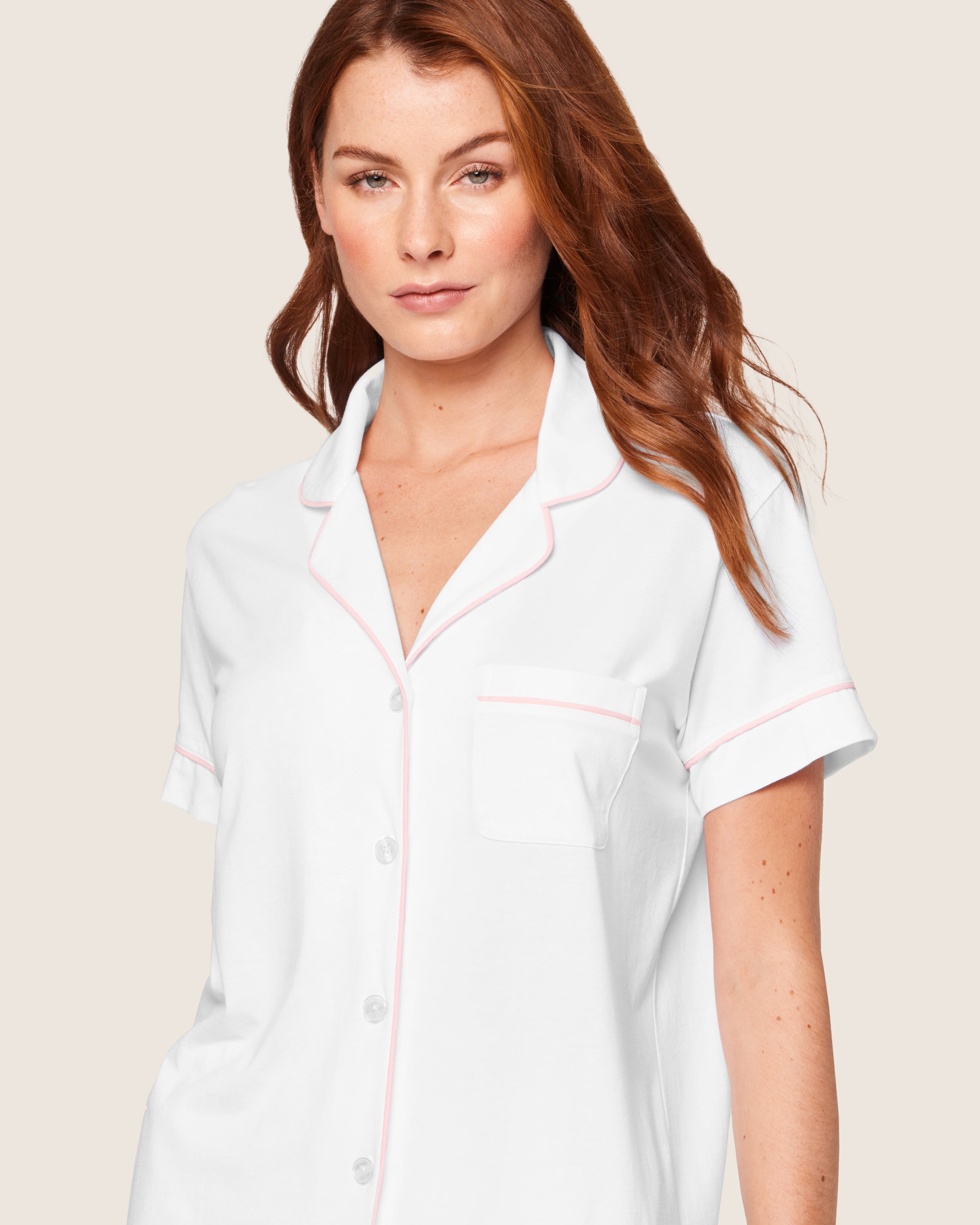 A person with long auburn hair wears a Petite Plume Womens Pima Pajama Short Set in White with Pink Piping. The luxurious cotton top features short sleeves and a chest pocket, set against a plain light beige background.