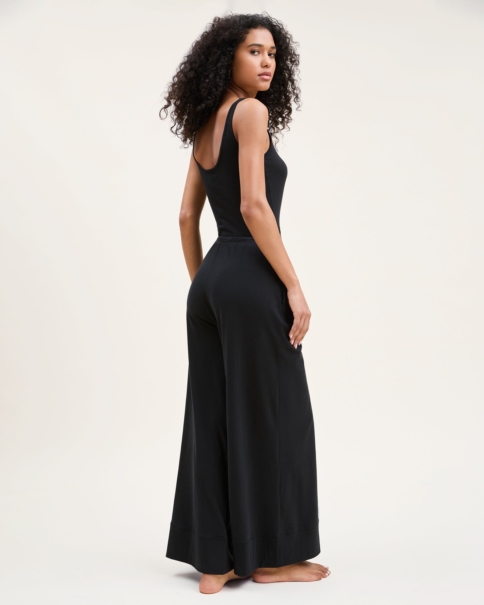 A woman with curly hair stands barefoot against a plain background, wearing the Women's Pima Wide-Leg Lounge Pant in Black and a sleeveless black tank top, looking relaxed and confident.