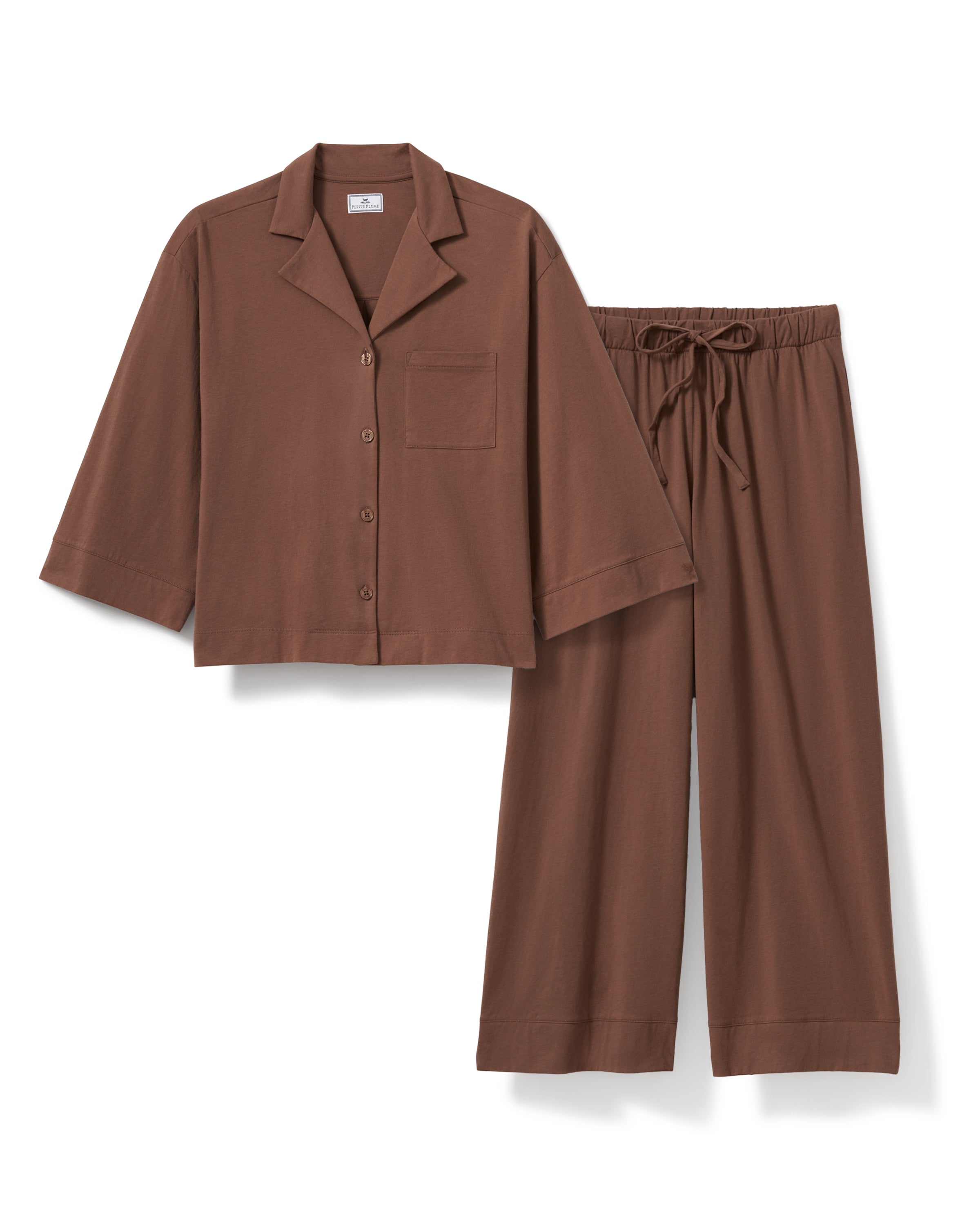 A woman with long curly hair stands barefoot against a plain background, wearing The Olivia Wide Leg Lightweight Pima Pajama Set in Chocolate Brown—a loose, button-down top and wide-leg pants—for a relaxed look.
