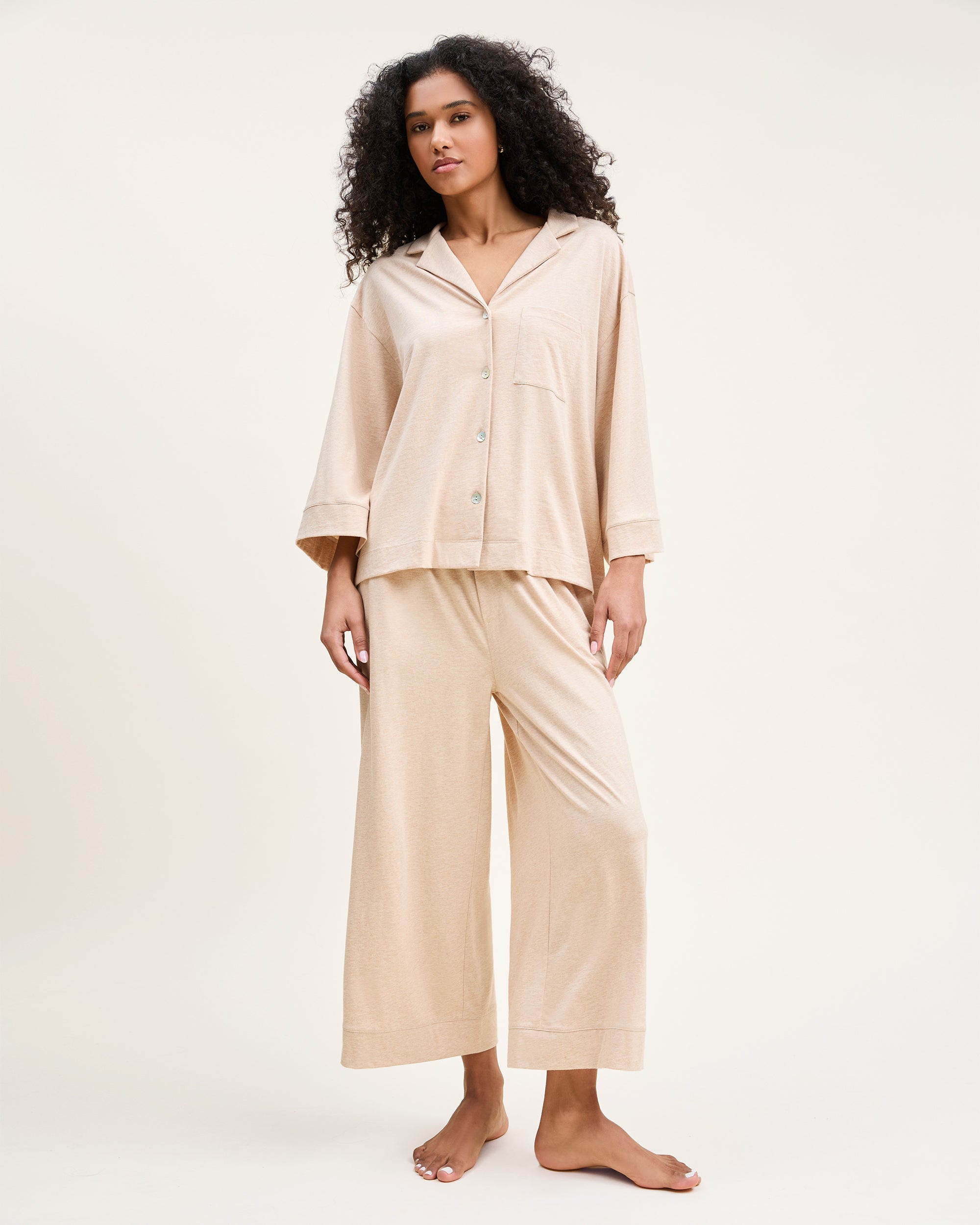 Against a plain backdrop, an individual with long curls and a relaxed stance wears The Olivia Wide Leg Pima Pajama Set in Camel by Petite Plume. The luxurious set features a button-up top and loose-fit pants, crafted from soft Pima cotton. They are barefoot, embodying comfort and elegance.