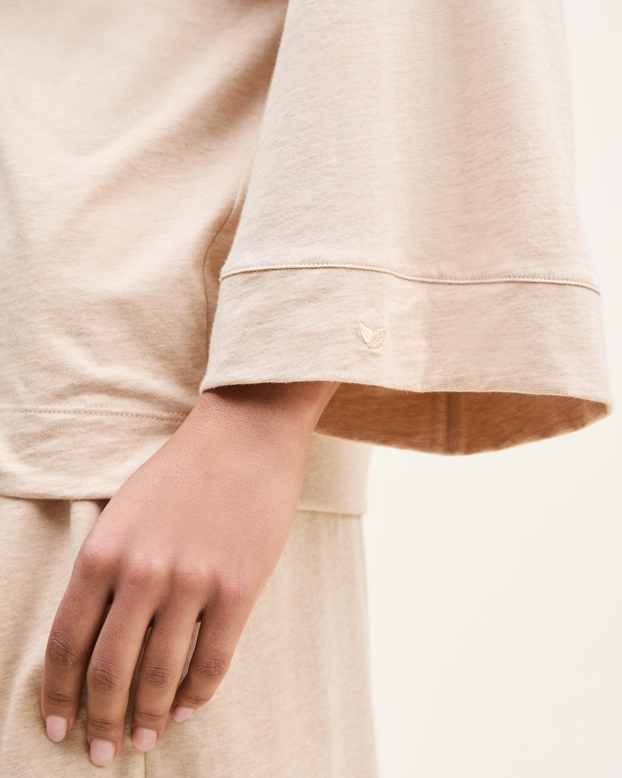 A close-up shows someone in The Olivia Wide Leg Pima Pajama Set in Camel by Petite Plume, highlighting the luxurious beige long-sleeve top with open cuffs and small embroidery. Their hand rests beside the matching pants, set against a soft neutral background.