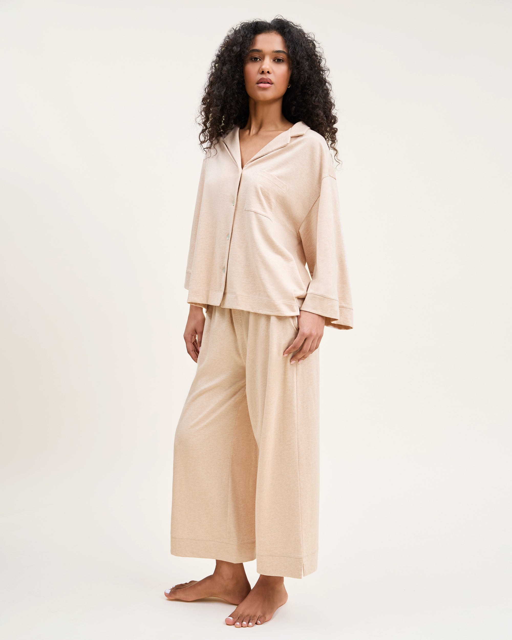 A person with long curly hair is standing barefoot, exuding luxury in Petite Plumes The Olivia Wide Leg Pima Pajama Set in Camel, featuring a beige buttoned top and wide-leg pants against a plain background.