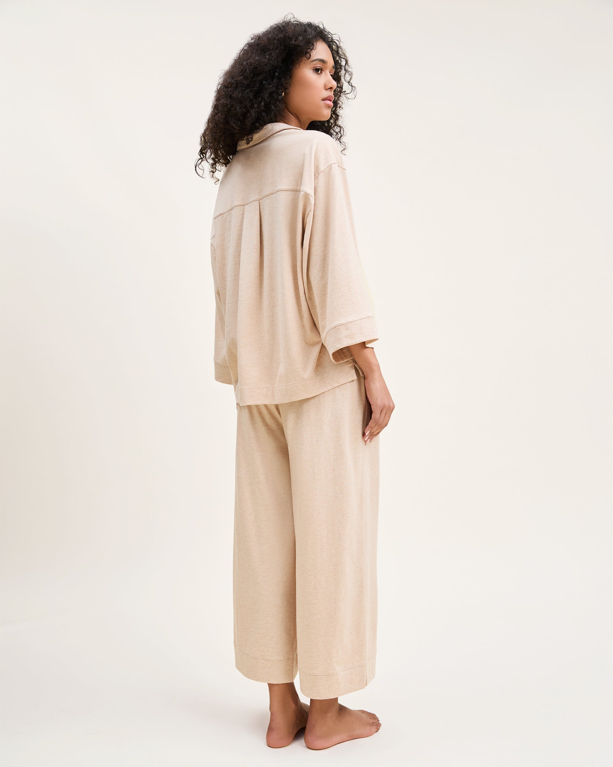 A curly-haired person stands barefoot against a neutral background, wearing The Olivia Wide Leg Pima Pajama Set in Camel by Petite Plume, crafted from luxurious Peruvian Pima cotton and looking to the side.