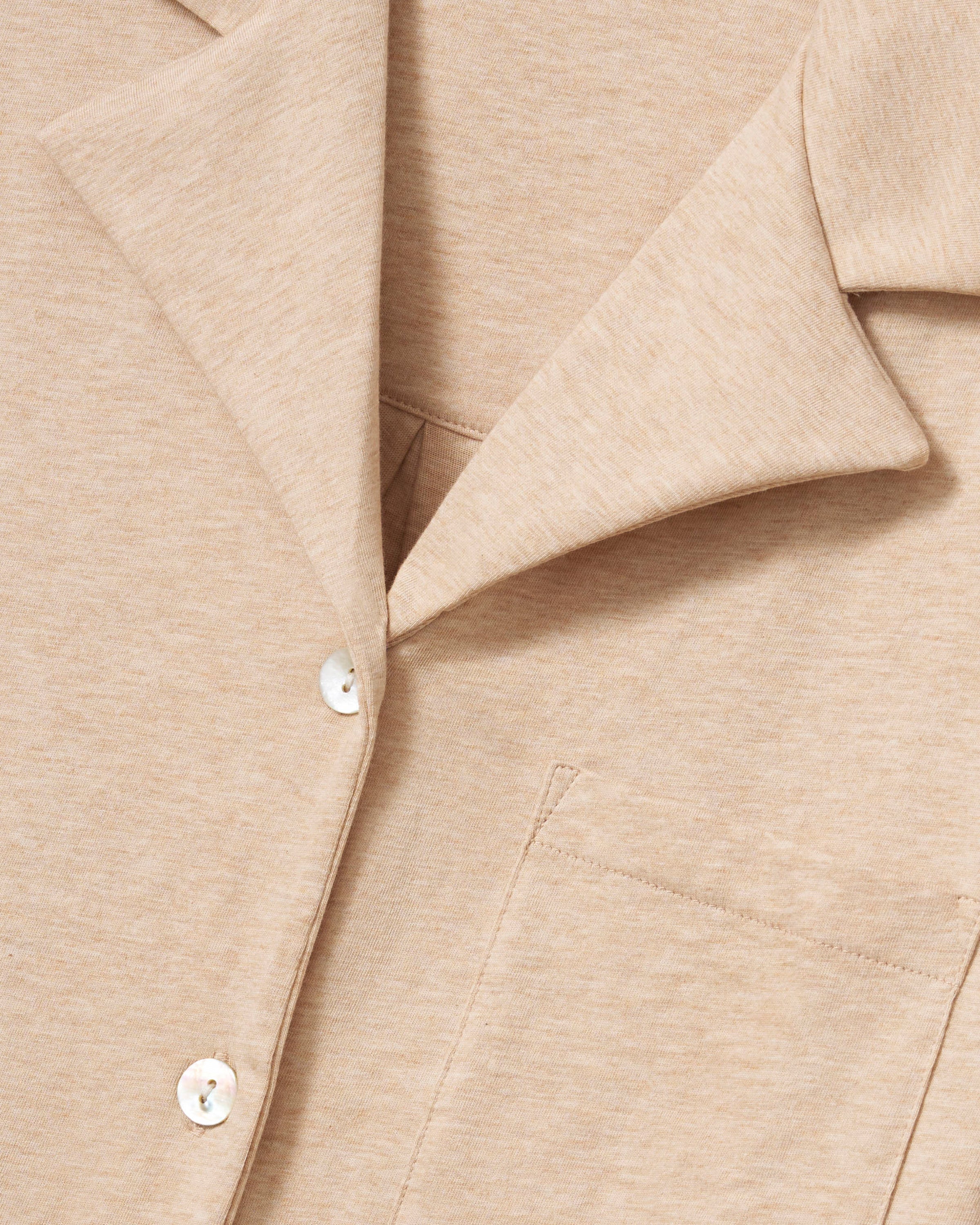 Against a plain backdrop, an individual with long curls and a relaxed stance wears The Olivia Wide Leg Pima Pajama Set in Camel by Petite Plume. The luxurious set features a button-up top and loose-fit pants, crafted from soft Pima cotton. They are barefoot, embodying comfort and elegance.