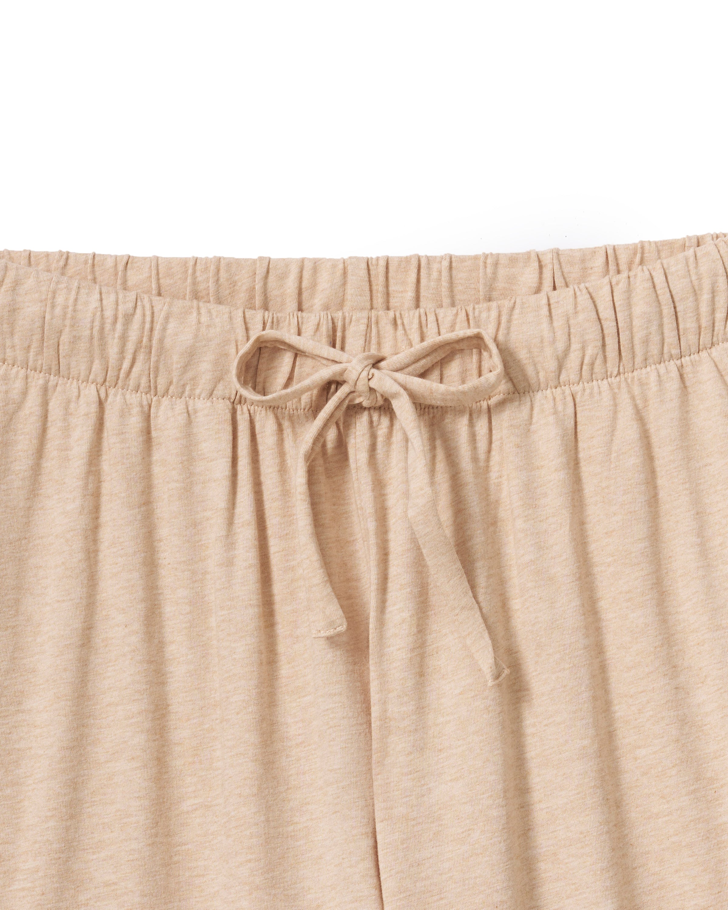 Against a plain backdrop, an individual with long curls and a relaxed stance wears The Olivia Wide Leg Pima Pajama Set in Camel by Petite Plume. The luxurious set features a button-up top and loose-fit pants, crafted from soft Pima cotton. They are barefoot, embodying comfort and elegance.