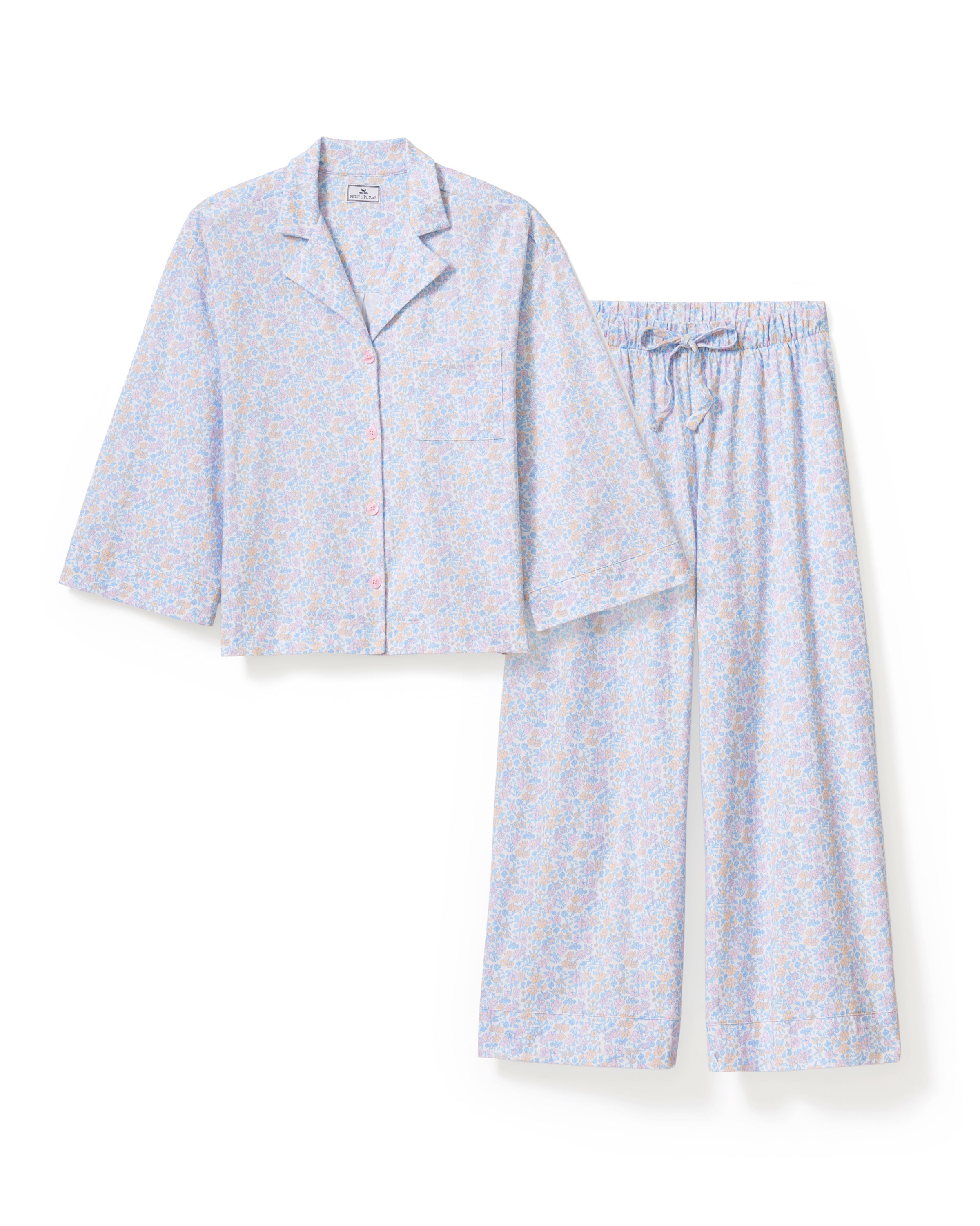 A woman stands barefoot against a plain background, wearing The Olivia Wide Leg Lightweight Pima Pajama Set in Fleur Blanche, featuring a loose long-sleeve button-up top and wide-leg pants with a light pastel floral pattern. She looks to the side and smiles.