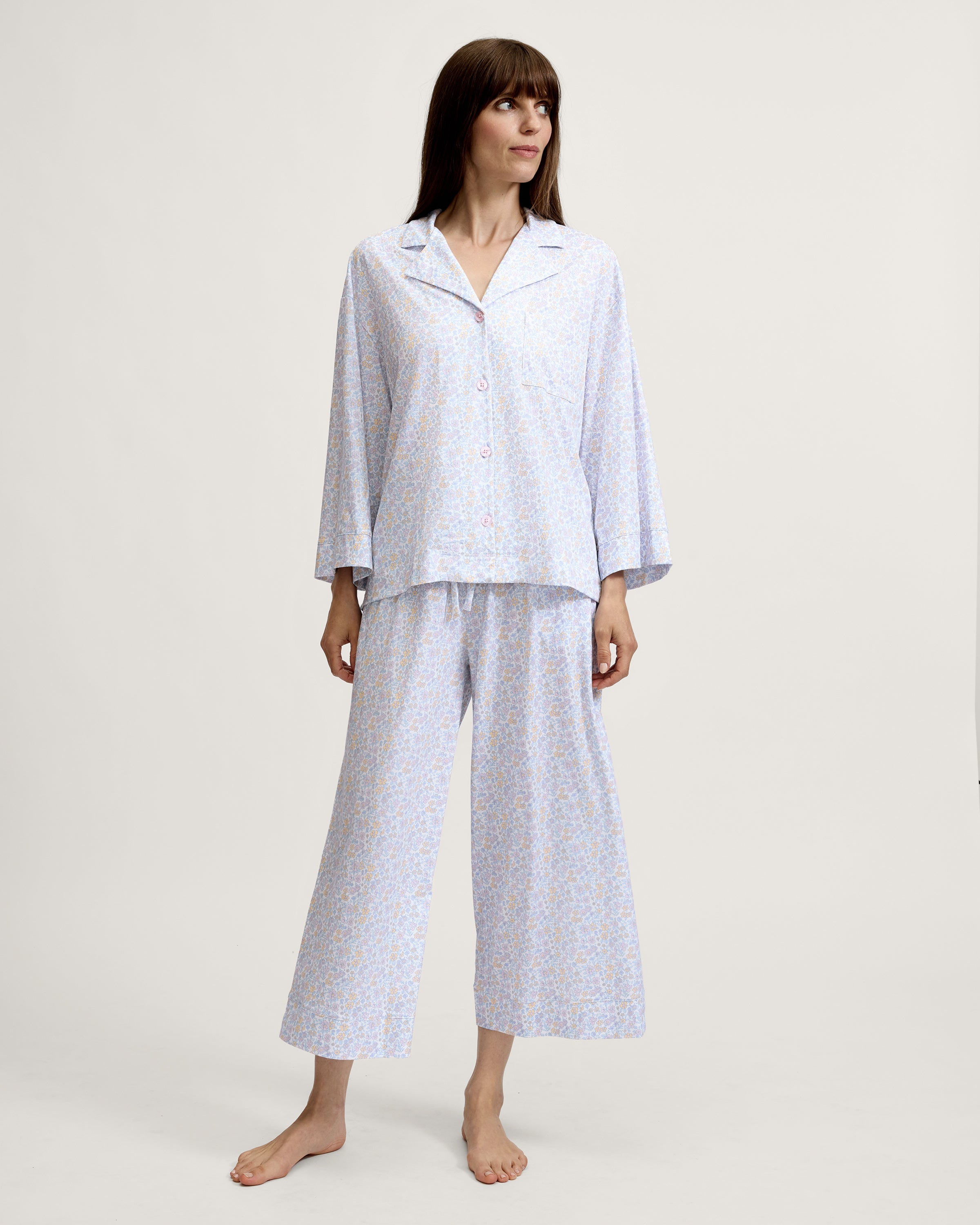 A woman stands barefoot against a plain background, wearing The Olivia Wide Leg Lightweight Pima Pajama Set in Fleur Blanche, featuring a loose long-sleeve button-up top and wide-leg pants with a light pastel floral pattern. She looks to the side and smiles.