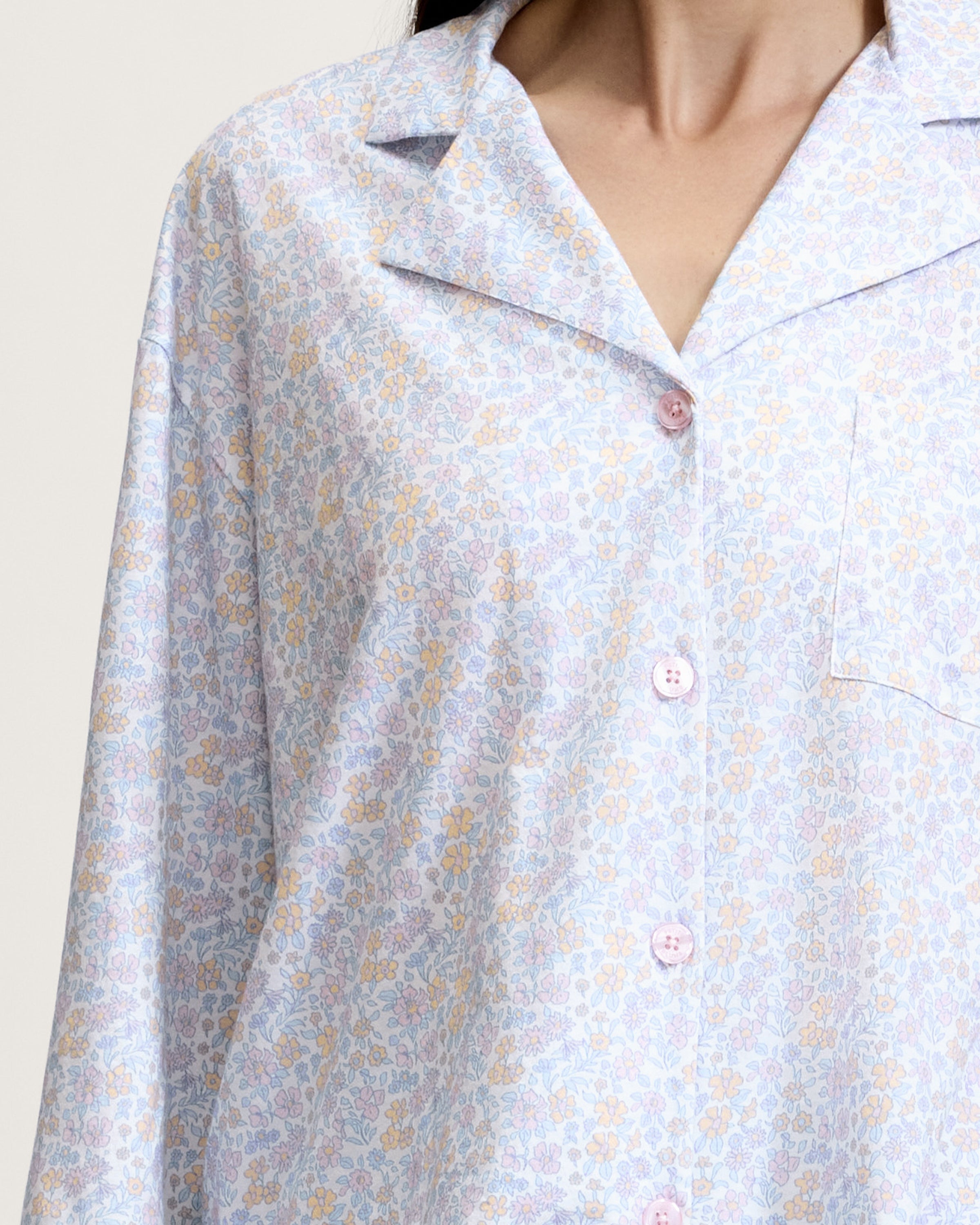 A woman stands barefoot against a plain background, wearing The Olivia Wide Leg Lightweight Pima Pajama Set in Fleur Blanche, featuring a loose long-sleeve button-up top and wide-leg pants with a light pastel floral pattern. She looks to the side and smiles.