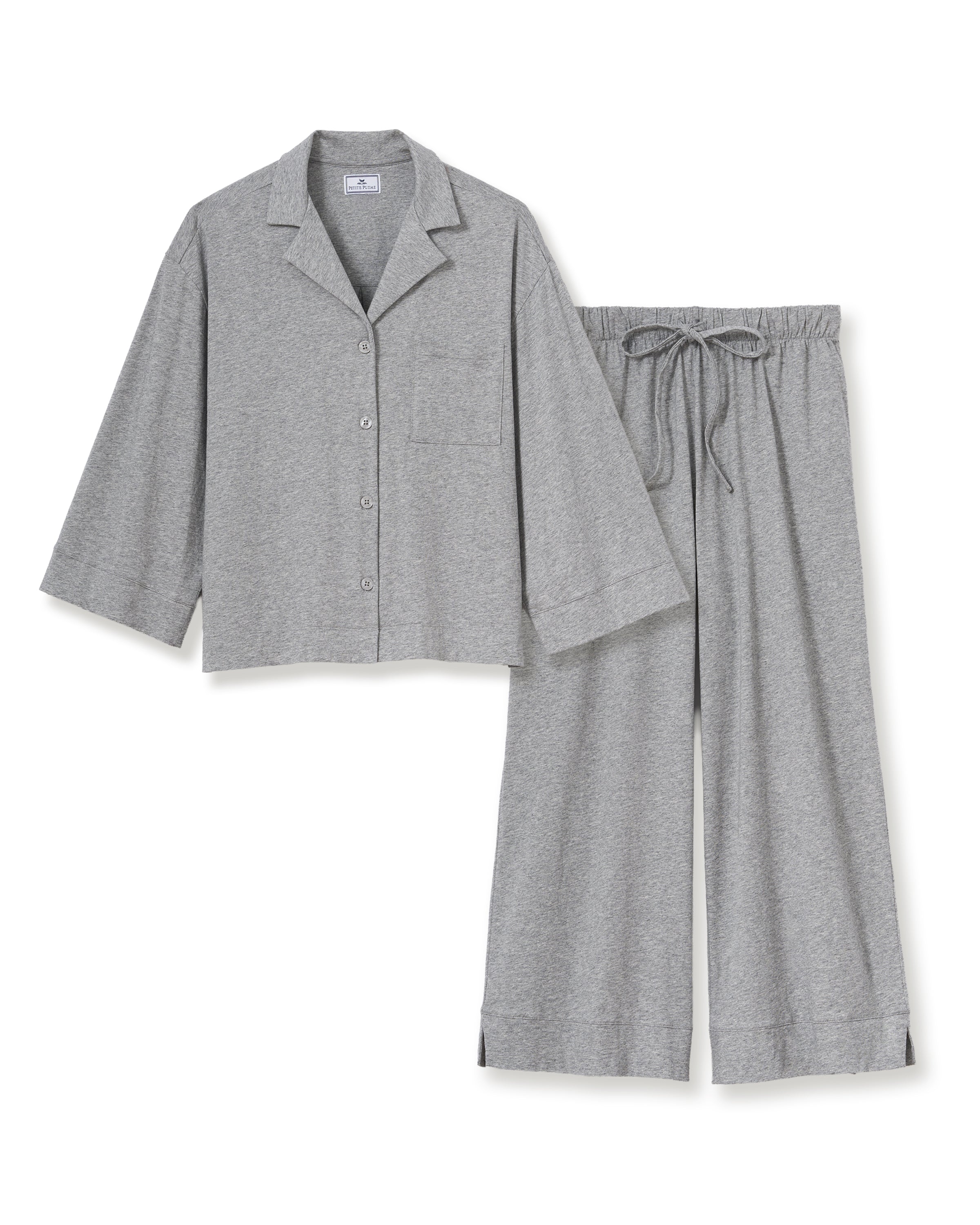 A woman stands barefoot against a plain background, smiling gently with one hand by her side, wearing The Olivia Wide Leg Lightweight Pima Pajama Set in Heather Grey—loose-fitting pants and a matching long-sleeve button-up top.