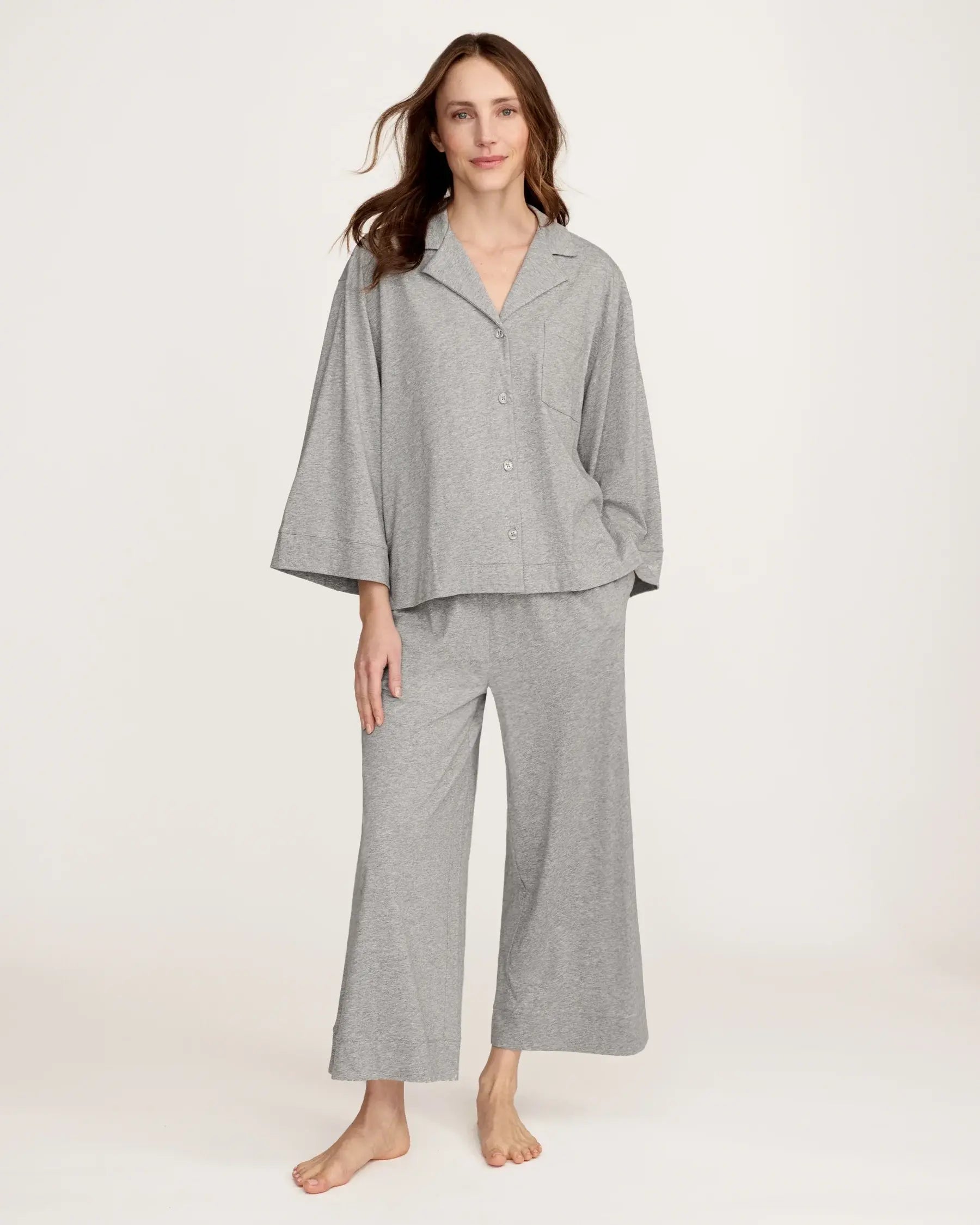 A woman stands barefoot against a plain background, smiling gently with one hand by her side, wearing The Olivia Wide Leg Lightweight Pima Pajama Set in Heather Grey—loose-fitting pants and a matching long-sleeve button-up top.