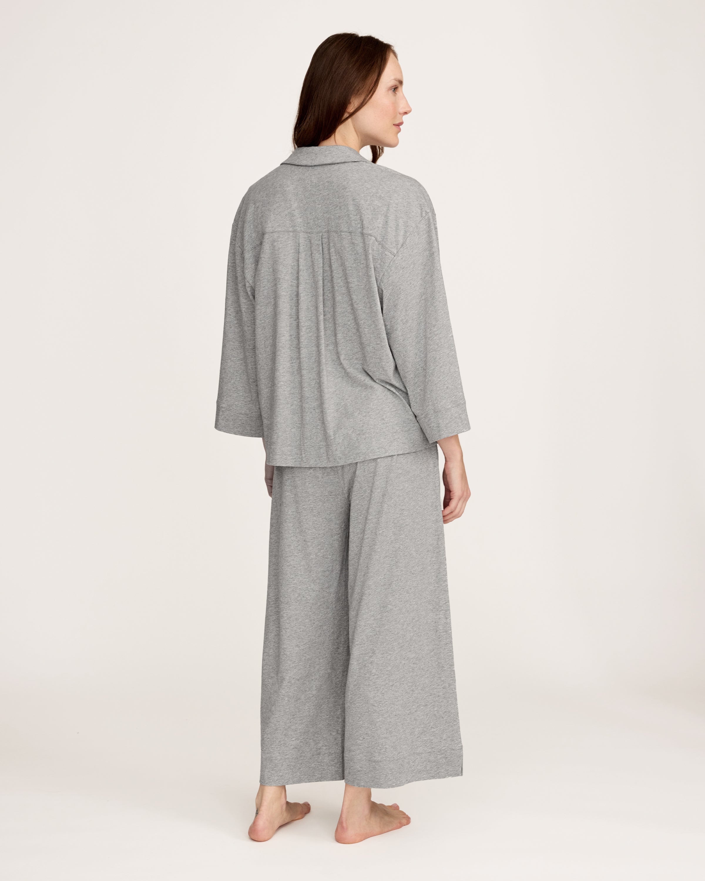 A woman stands barefoot against a plain background, smiling gently with one hand by her side, wearing The Olivia Wide Leg Lightweight Pima Pajama Set in Heather Grey—loose-fitting pants and a matching long-sleeve button-up top.
