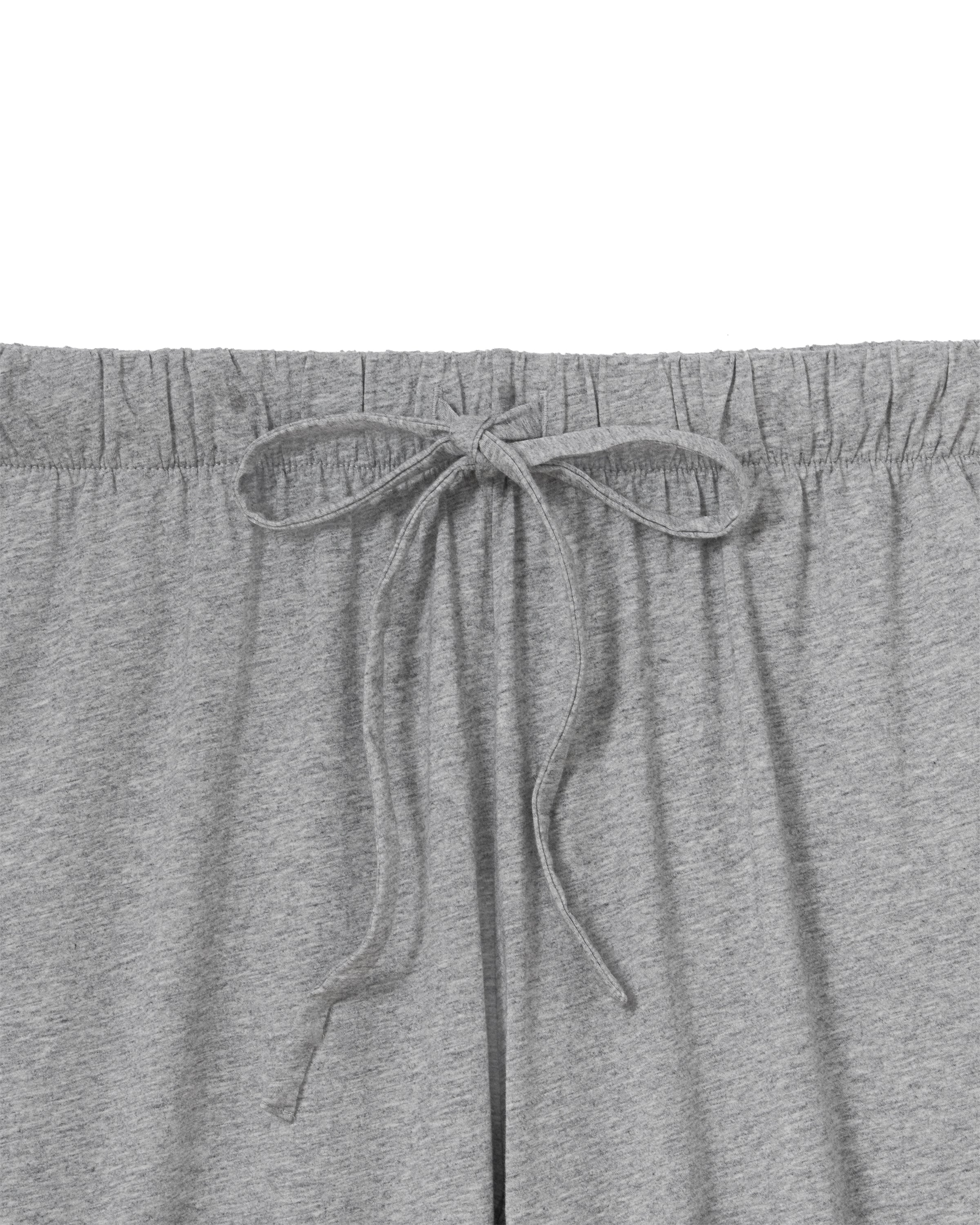 A woman stands barefoot against a plain background, smiling gently with one hand by her side, wearing The Olivia Wide Leg Lightweight Pima Pajama Set in Heather Grey—loose-fitting pants and a matching long-sleeve button-up top.