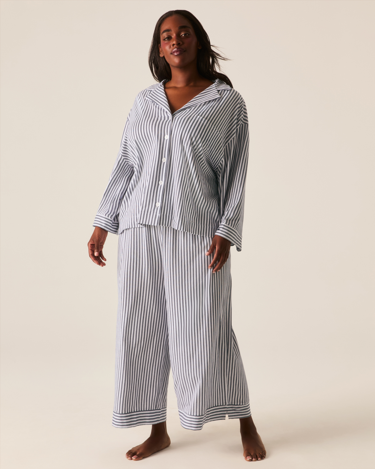A person wears The Olivia Wide Leg Pima Pajama Set in Navy French Ticking, featuring a navy and white striped button-up long-sleeved top and wide-leg trousers.