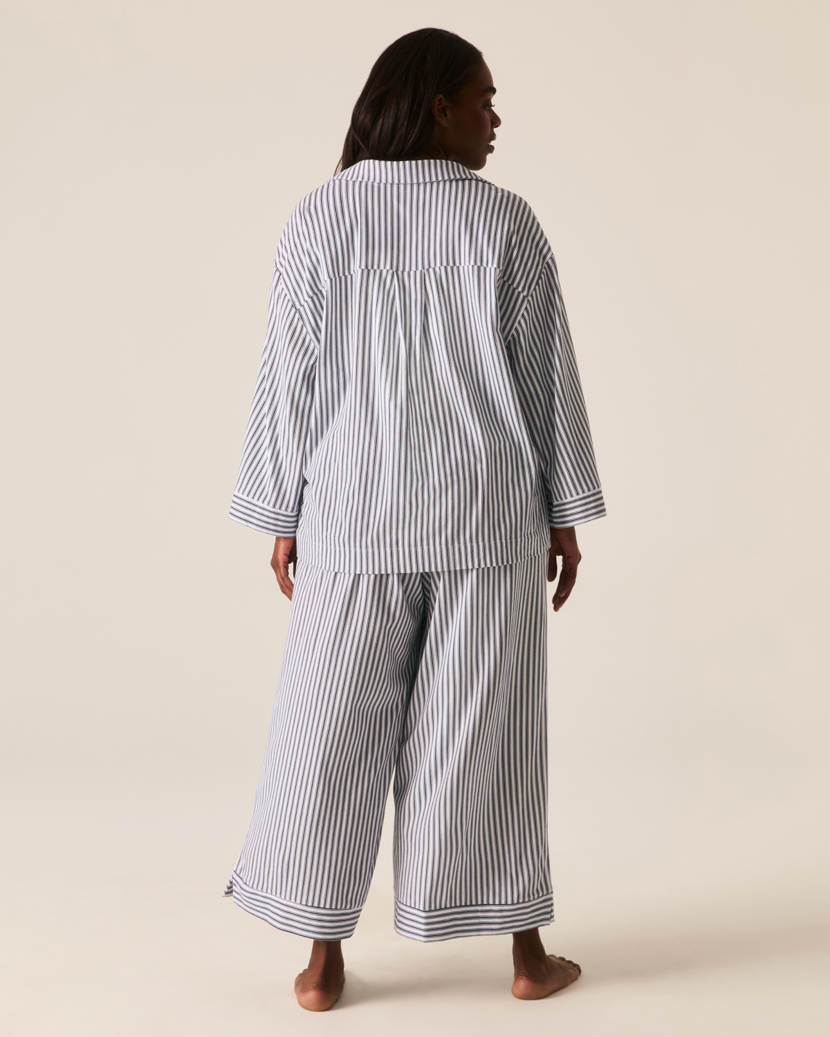 A person seen from behind wears The Olivia Wide Leg Pima Pajama Set in Navy French Ticking, featuring a long-sleeved button-down shirt and wide-leg trousers in a blue and white striped pattern.
