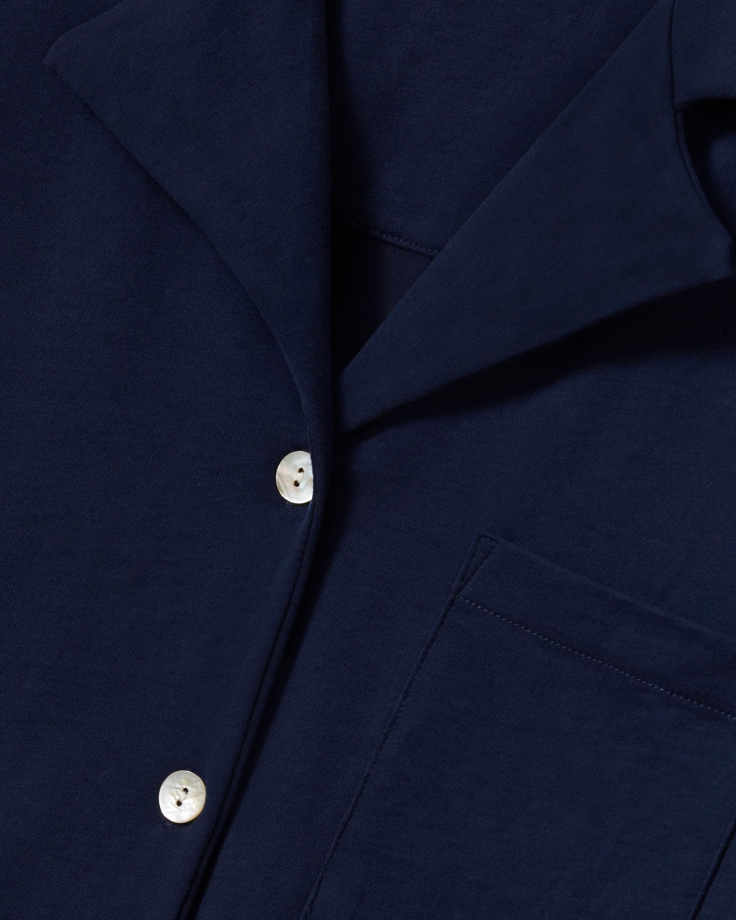 A person with long brown hair wears The Olivia Wide Leg Pima Pajama Set in Navy by Petite Plume, featuring a luxurious, buttoned, long-sleeved top and elegant wide-legged pants. They stand barefoot against an off-white background.