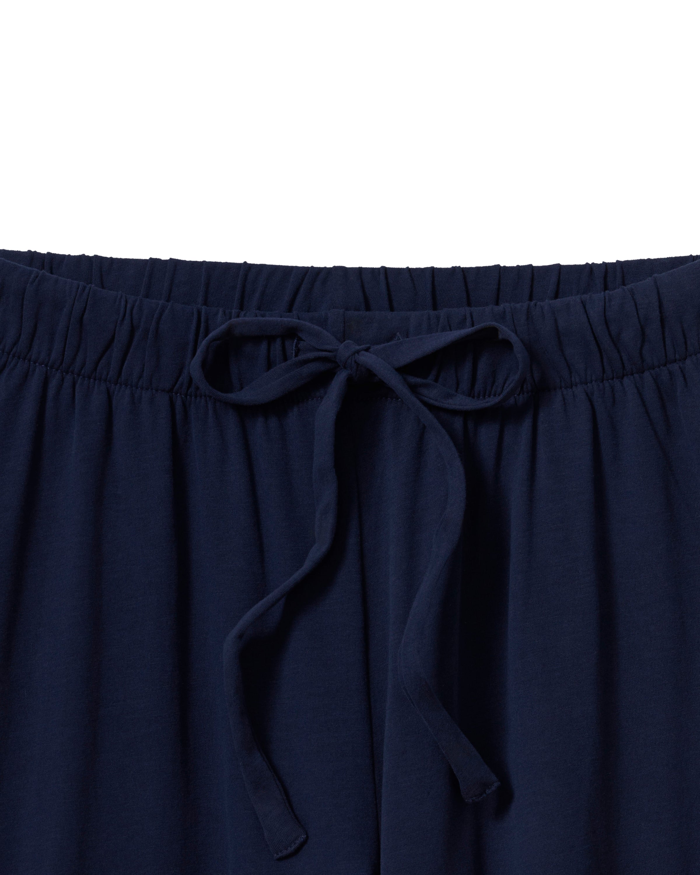 A person with long brown hair wears The Olivia Wide Leg Pima Pajama Set in Navy by Petite Plume, featuring a luxurious, buttoned, long-sleeved top and elegant wide-legged pants. They stand barefoot against an off-white background.