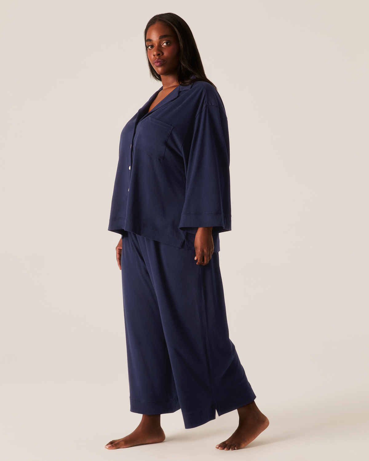 A person stands wearing The Olivia Wide Leg Pima Pajama Set in Navy, featuring a long-sleeved button-down shirt and wide-leg trousers.