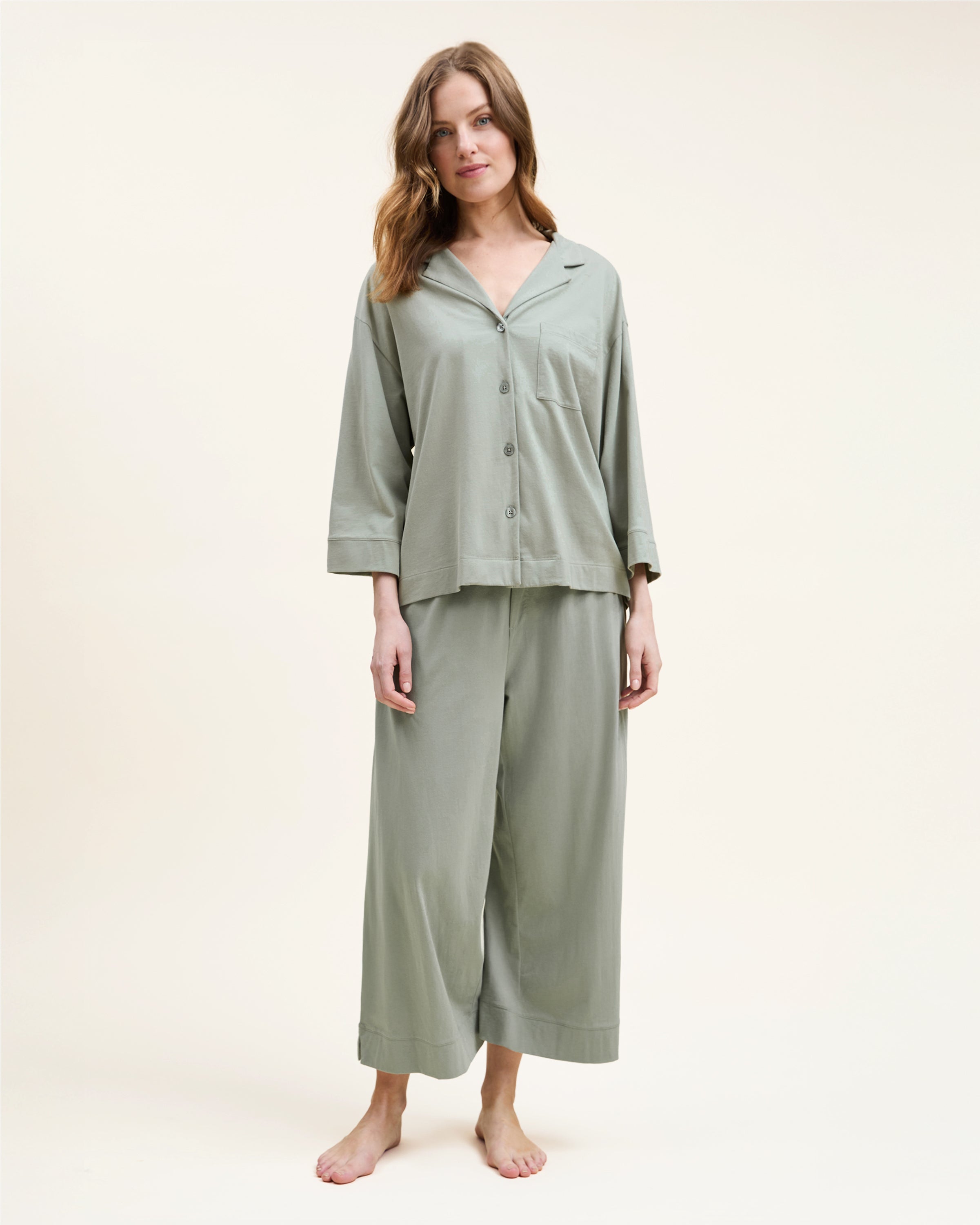 The Olivia Wide Leg Pima Pajama Set in Sage includes a button-up, collared top with a chest pocket and three-quarter sleeves, plus matching elastic waist wide-leg pants. Both pieces are neatly laid flat on a white background.