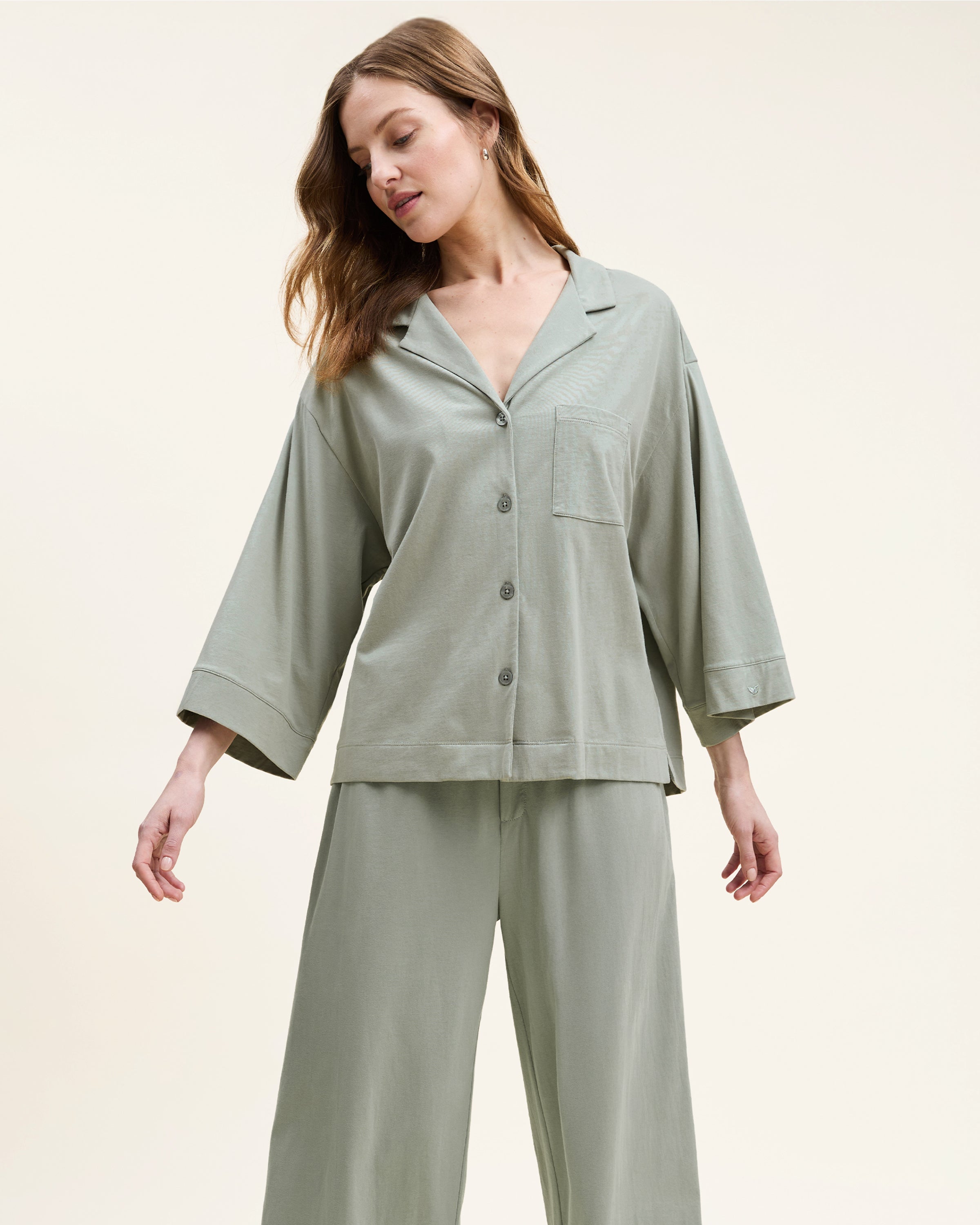 The Olivia Wide Leg Pima Pajama Set in Sage includes a button-up, collared top with a chest pocket and three-quarter sleeves, plus matching elastic waist wide-leg pants. Both pieces are neatly laid flat on a white background.