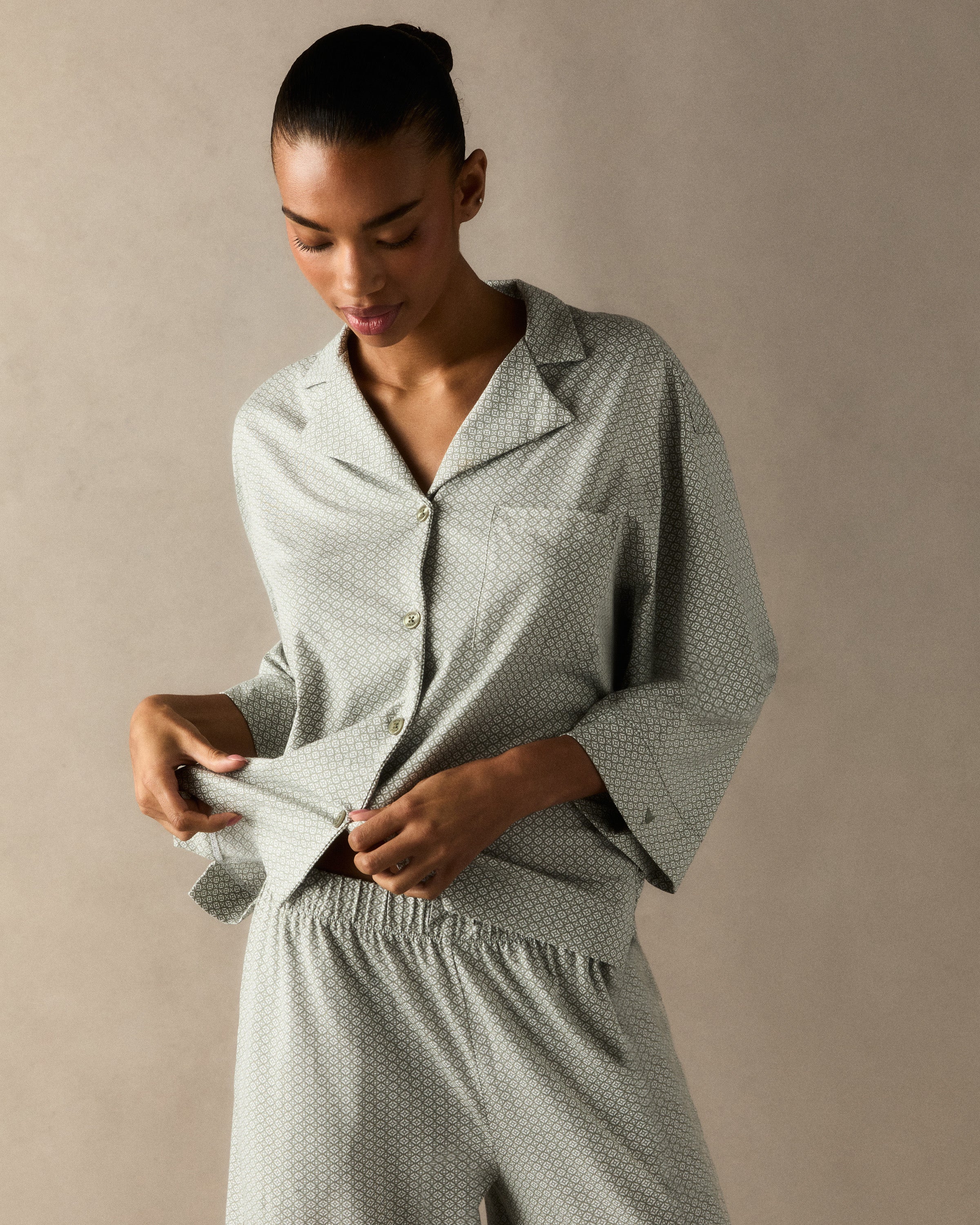 A woman stands barefoot against a plain white background, wearing The Olivia Wide Leg Lightweight Pima Pajama Set in Sage Geo—a matching loose-fitting ensemble with long sleeves and wide-leg pants in a subtle light pattern.