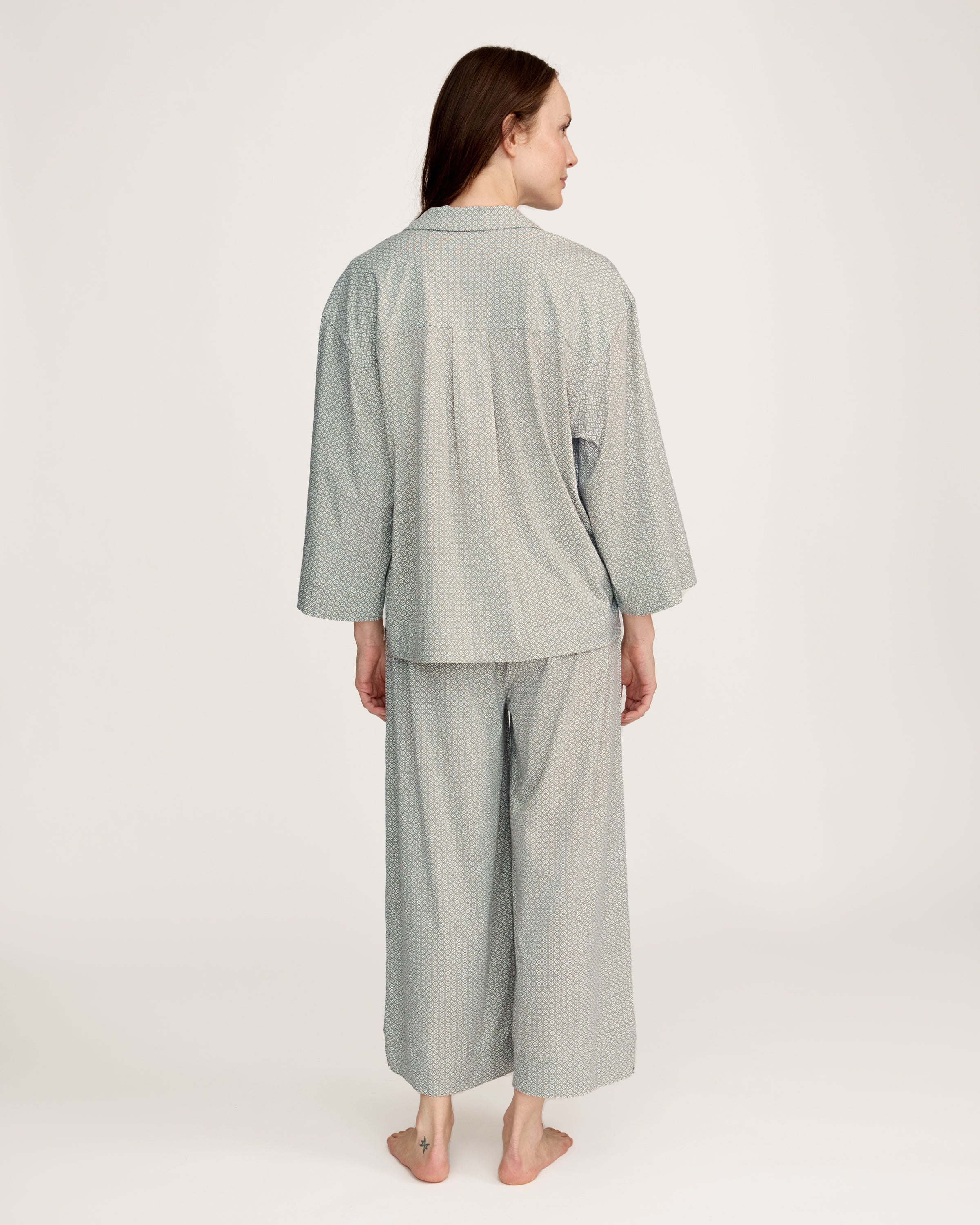 A woman stands barefoot against a plain white background, wearing The Olivia Wide Leg Lightweight Pima Pajama Set in Sage Geo—a matching loose-fitting ensemble with long sleeves and wide-leg pants in a subtle light pattern.