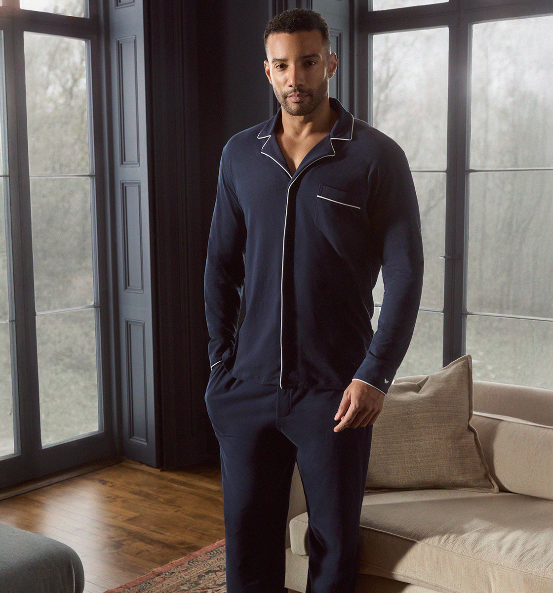 A man wearing navy blue pajamas with white piping stands in a stylish living room with large windows, a beige sofa, and wooden floors.