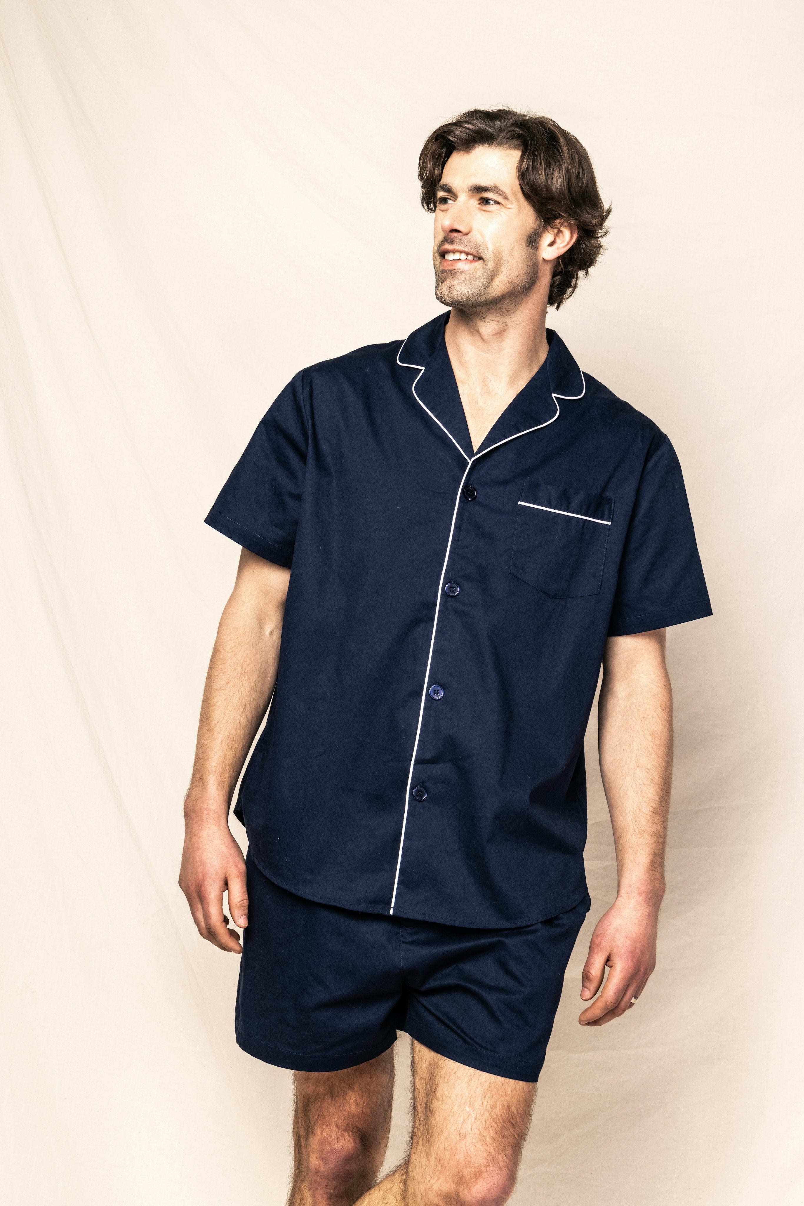 A man is wearing the Mens Twill Pajama Short Set in Navy by Petite Plume with white piping. He stands against a light background, looking to the side and smiling, one hand down and the other slightly raised.