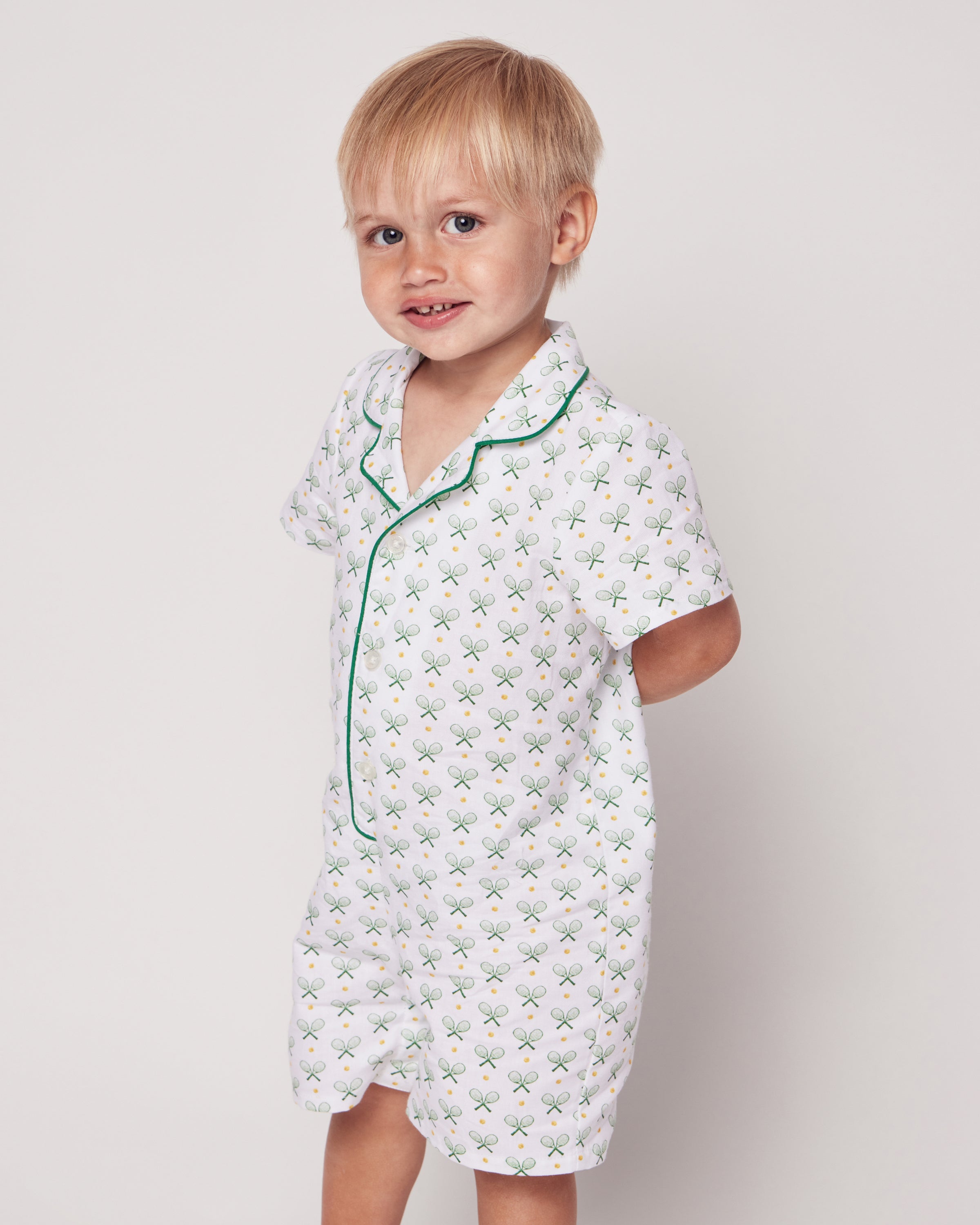 A young child with blonde hair wears the luxurious cotton Babys Twill Summer Romper in Match Point by Petite Plume, featuring green tennis racket patterns and green piping. The child is smiling with hands behind their back against a plain backdrop.