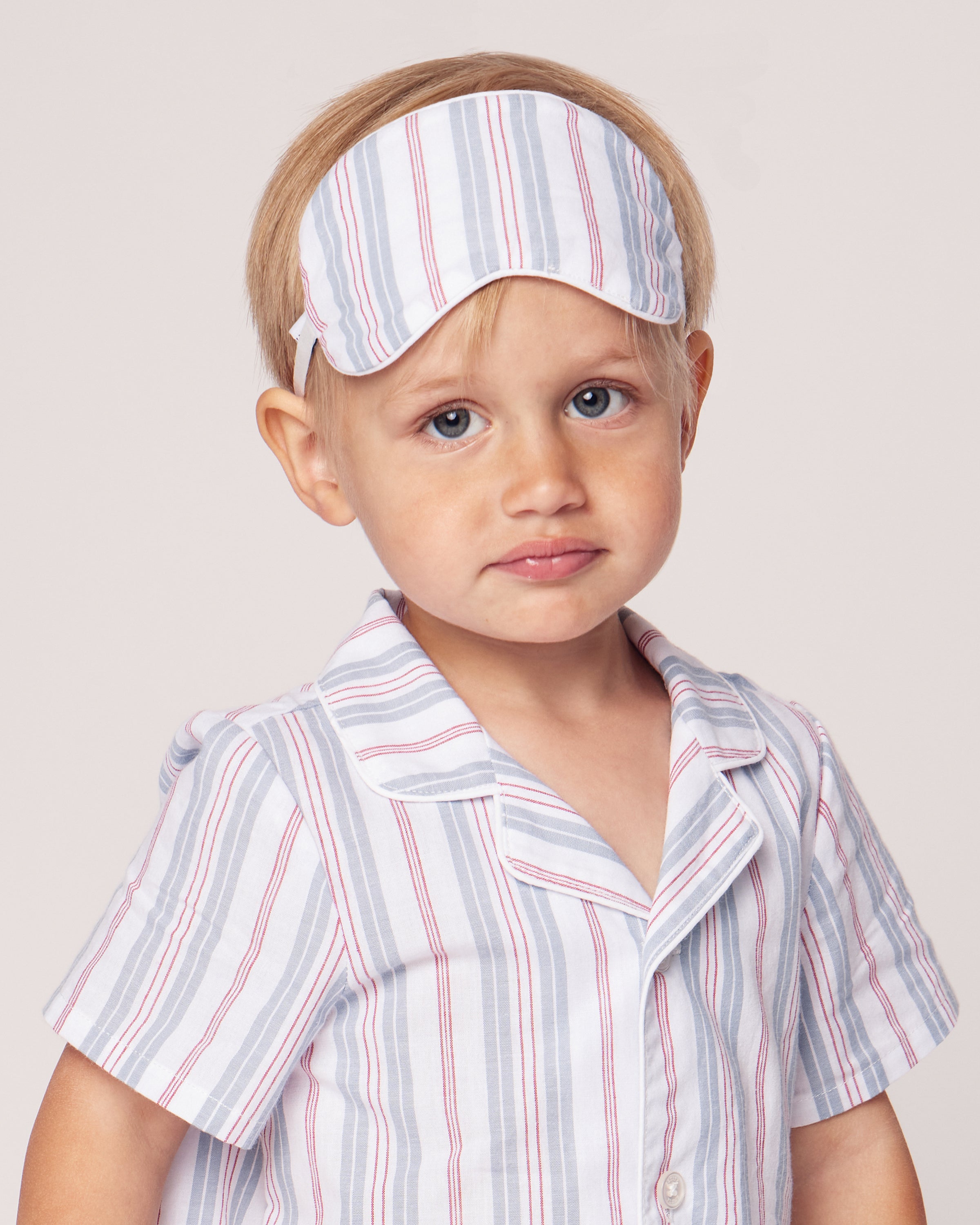 A young blond child dons a Kids Sleep Mask in Vintage French Stripes by Petite Plume, paired with a matching pajama shirt of blue and red vertical stripes on white. With a neutral expression against a plain backdrop, the scene evokes vintage charm.
