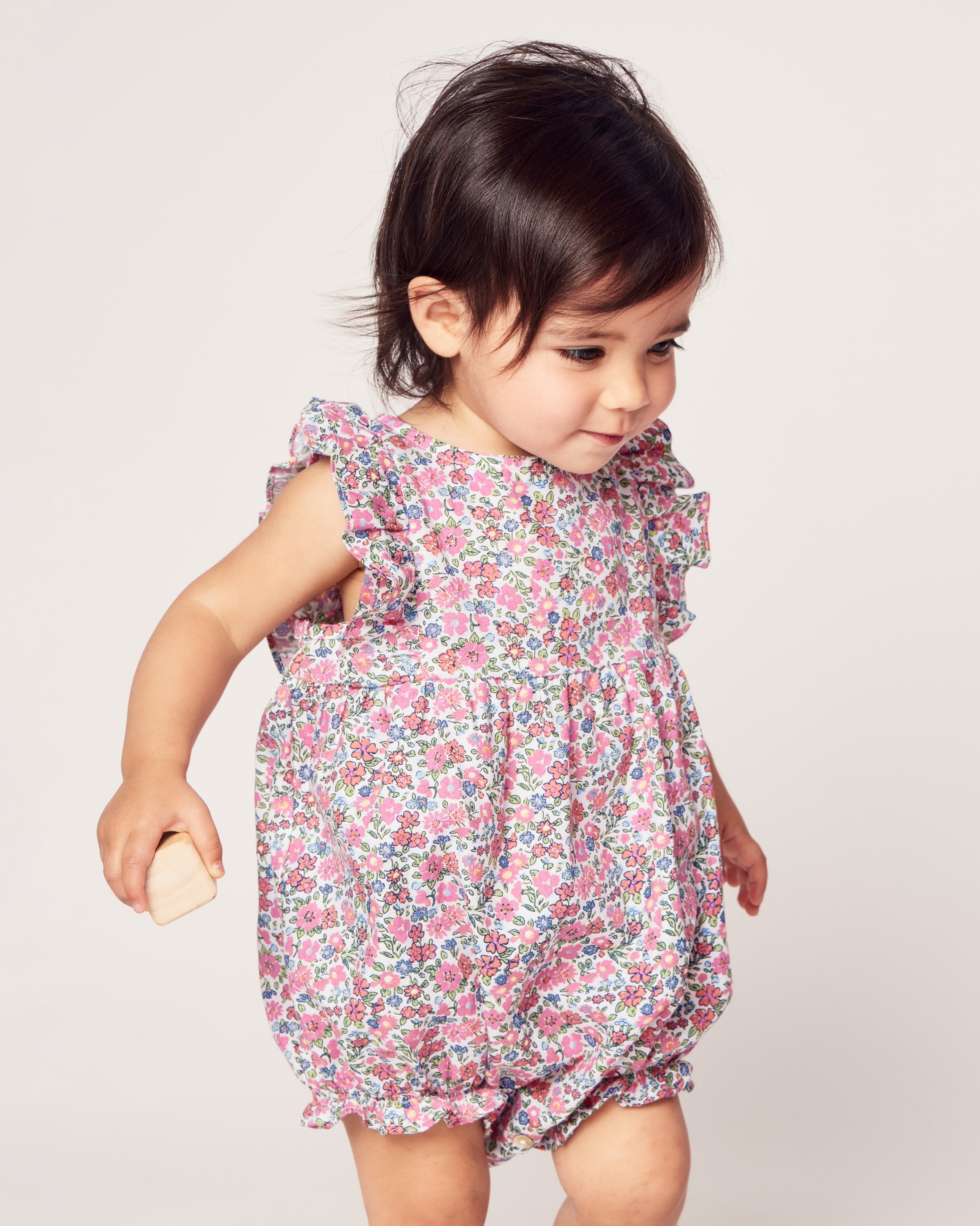 A toddler in Petite Plumes Babys Twill Ruffled Romper in Fleurs de Rose stands against a plain background, holding a wooden block. The ruffled sleeves of high-quality cotton enhance the charm as the child with short dark hair gazes down slightly.