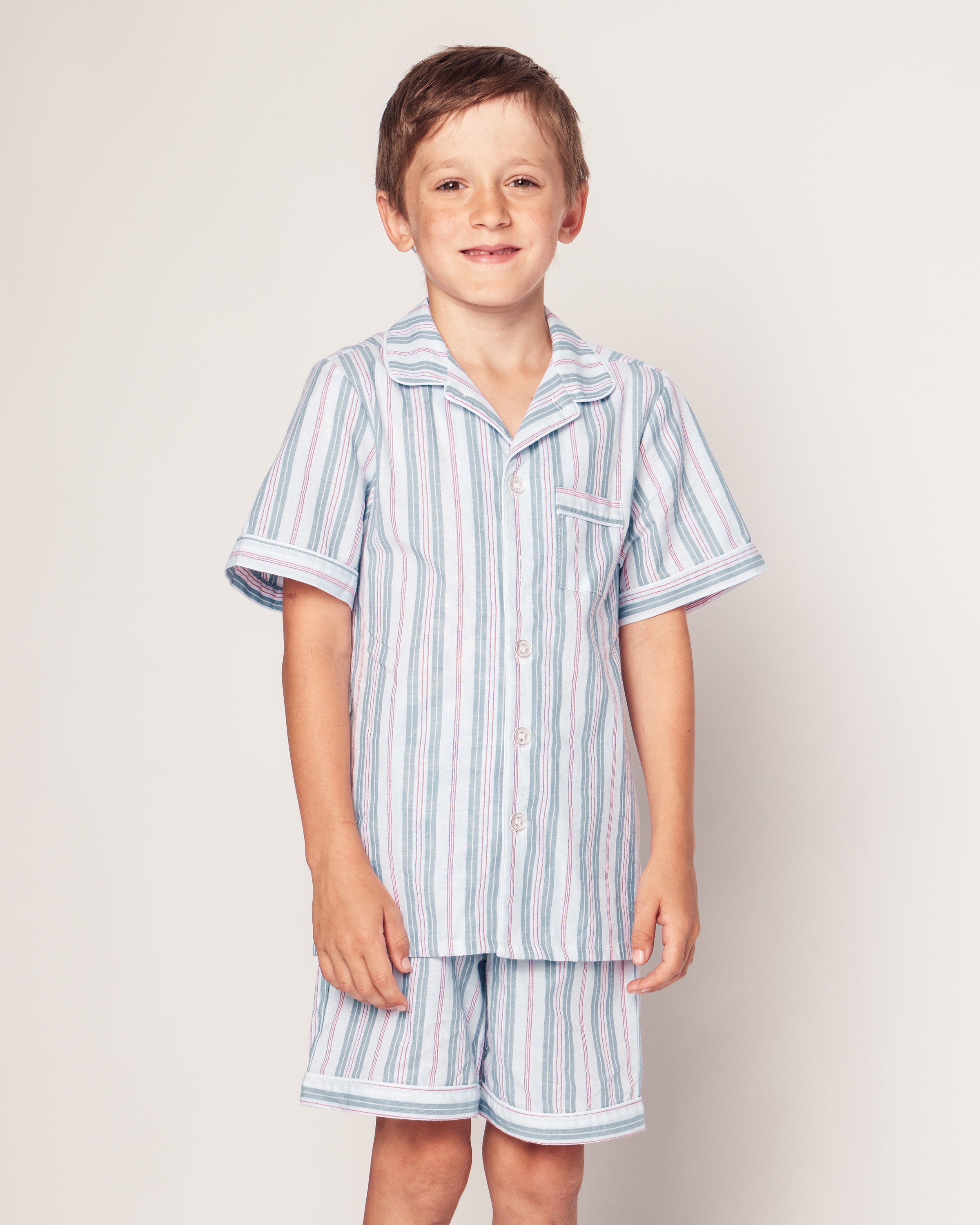 A young child smiles in a Petite Plume Kids Twill Pajama Short Set featuring a vintage French striped pattern in blue, white, and pink with a chest pocket. They stand against a plain background.