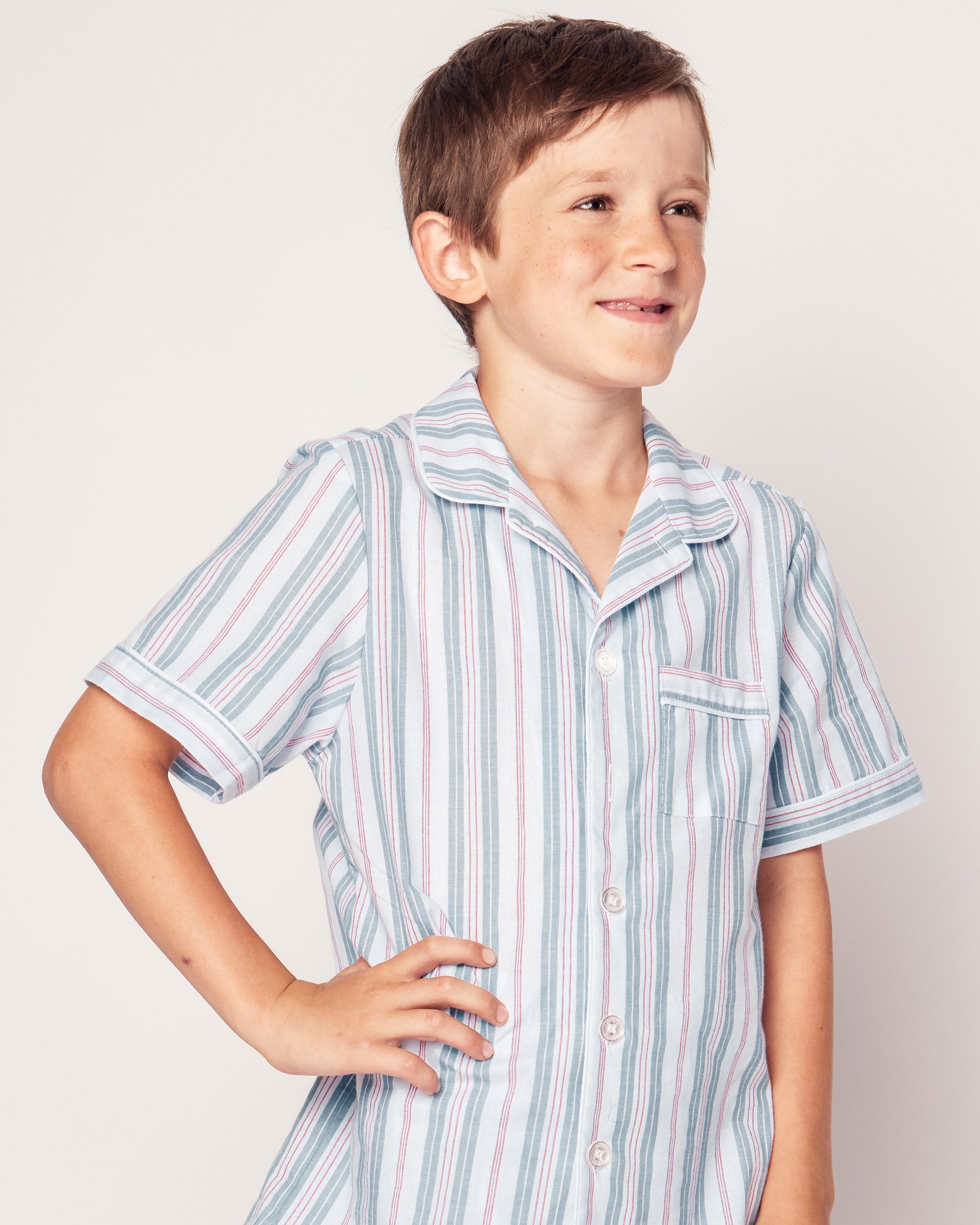 A young boy with short brown hair smiles as he looks to the side, wearing Petite Plumes Kids Twill Pajama Short Set in Vintage French Stripes. His right hand rests on his hip against a plain background.