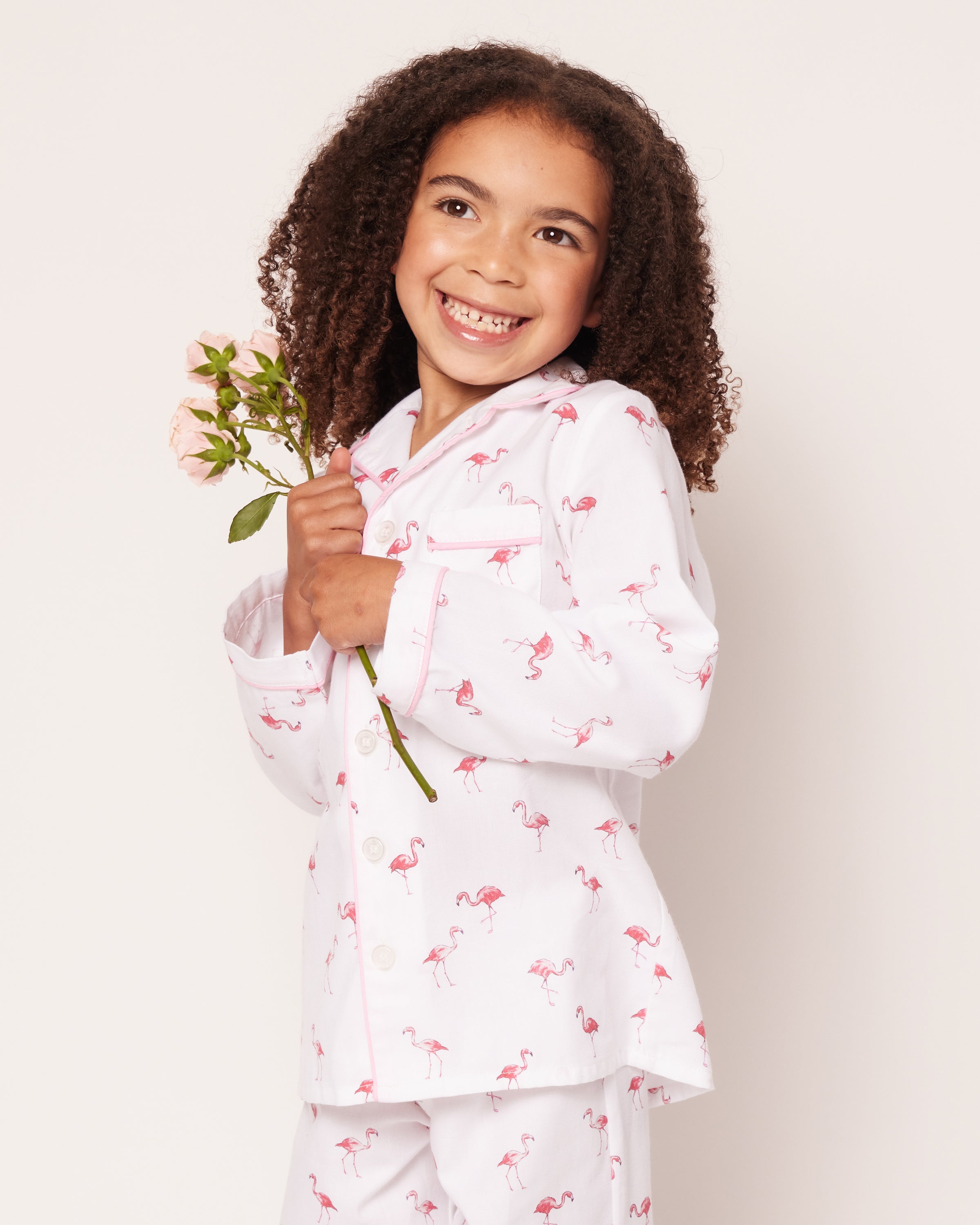 A young girl with curly hair smiles, holding pink roses while wearing the Kids Twill Pajama Set in Flamingos by Petite Plume. Her flame-retardant white pajamas ensure comfort and safety. The plain white background highlights her cheerful expression.