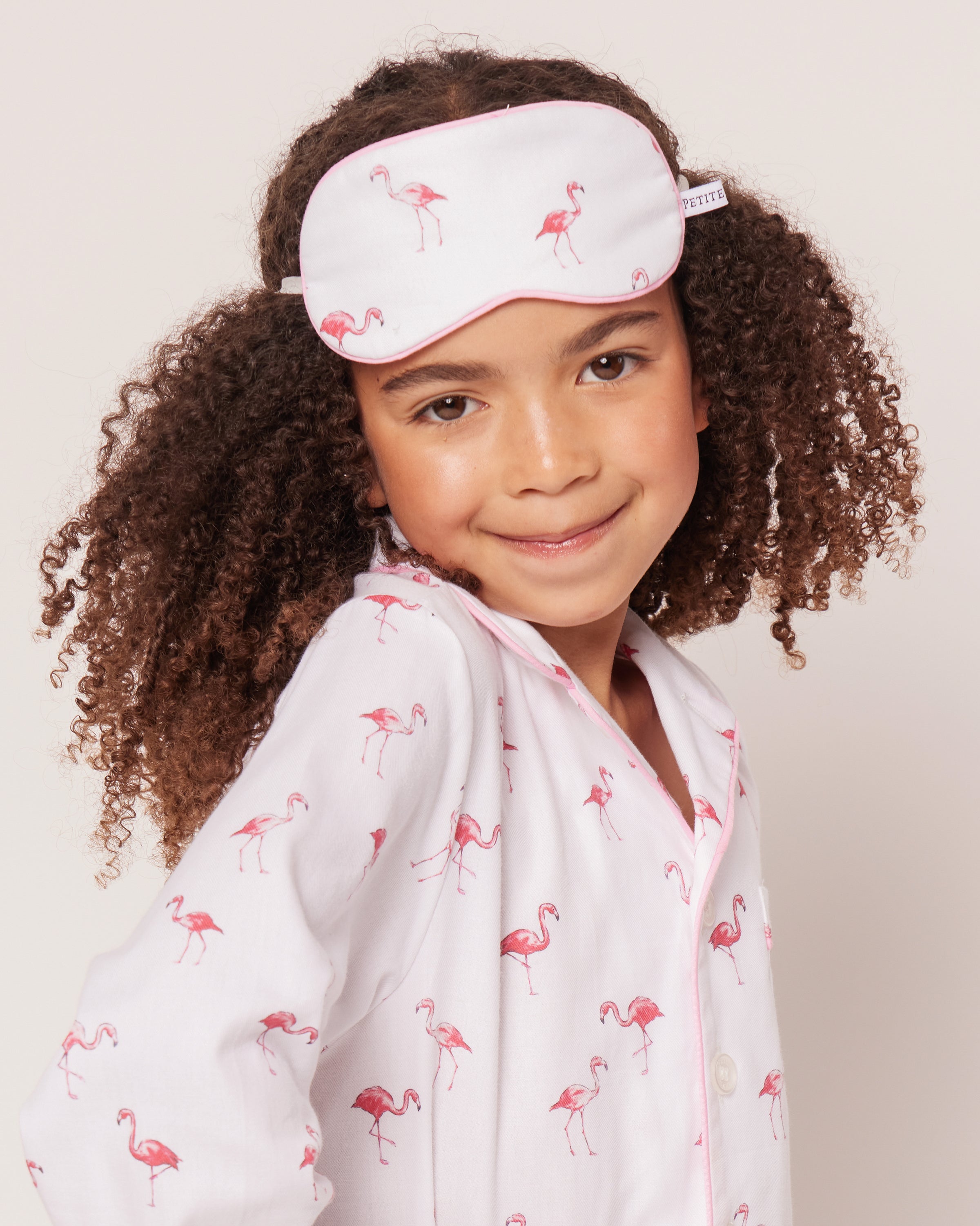 A smiling child with curly hair wears Petite Plumes Kids Sleep Mask in Flamingos and matching pajamas, delighting in their tropical print.