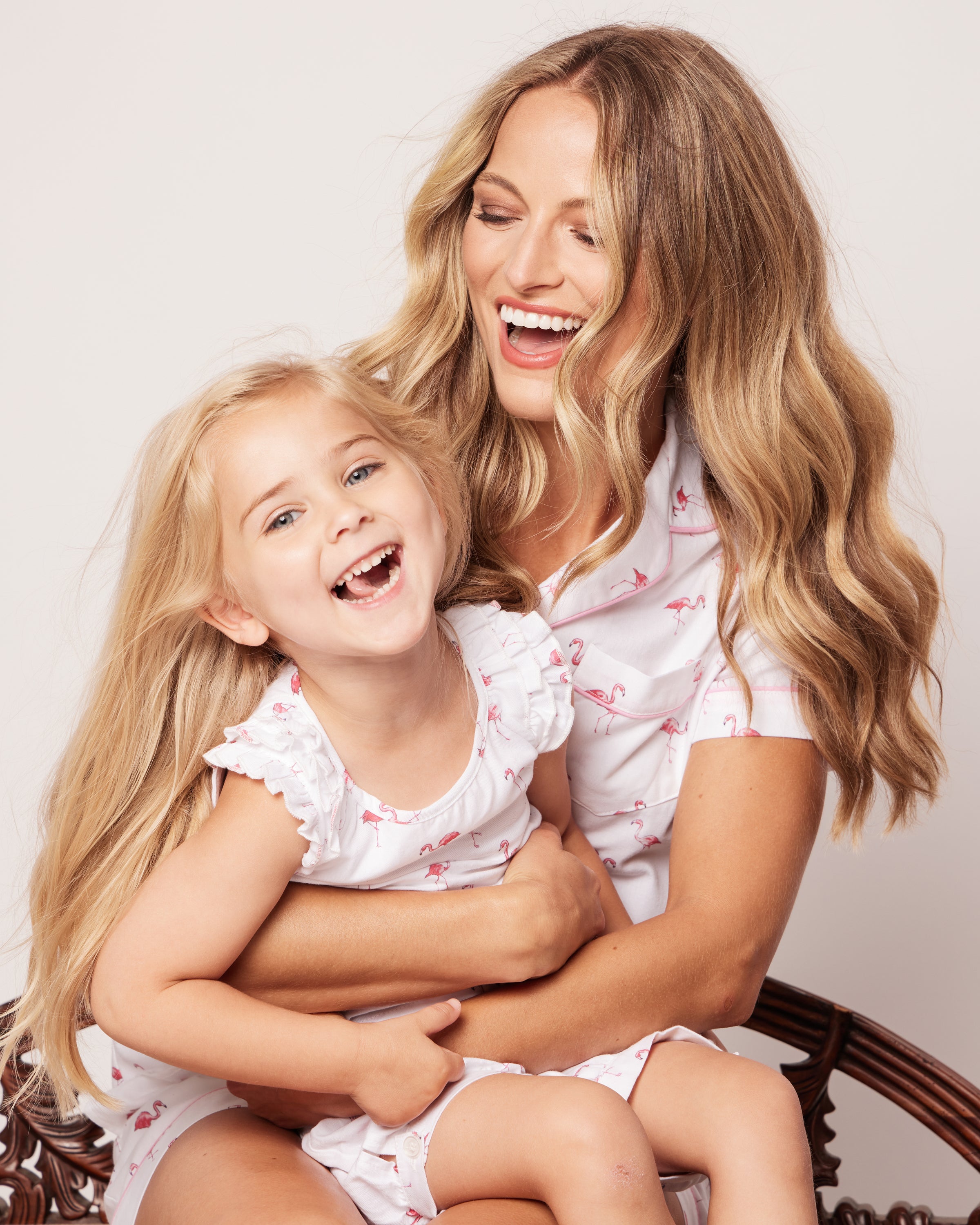 A woman and a young girl with long blonde hair are joyfully sitting together, wearing matching Petite Plume sleepwear. The girl is in the Girls Twill Amelie Short Set in Flamingos as they share a happy moment, the woman holding her close.