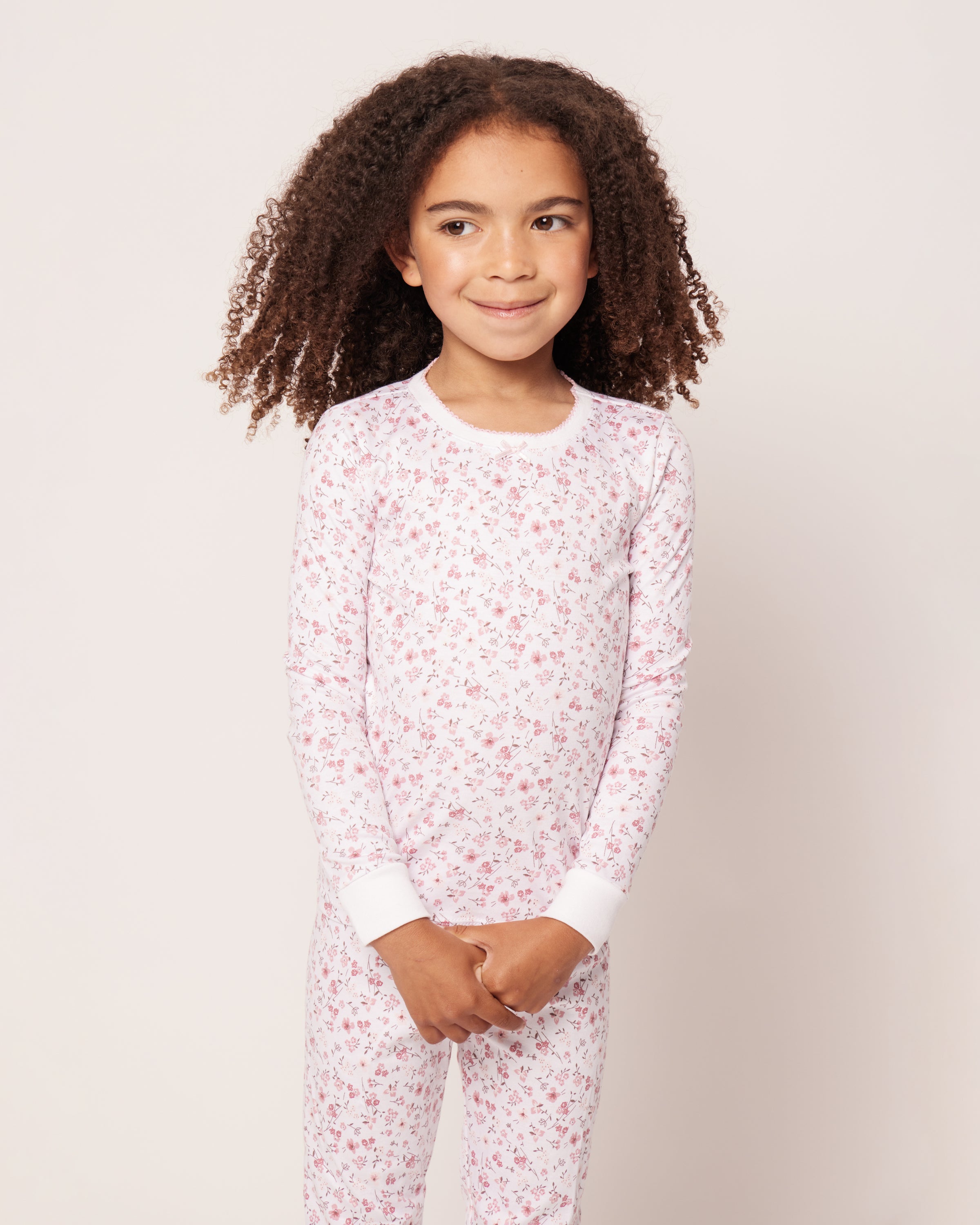 A curly-haired child beams in joy, wearing Petite Plumes Kids Pima Snug Fit Pajama Set in Dorset Floral. The soft, pink and white floral sleepwear includes a charming long-sleeve top and pants, as the child clasps their hands happily against a neutral backdrop.